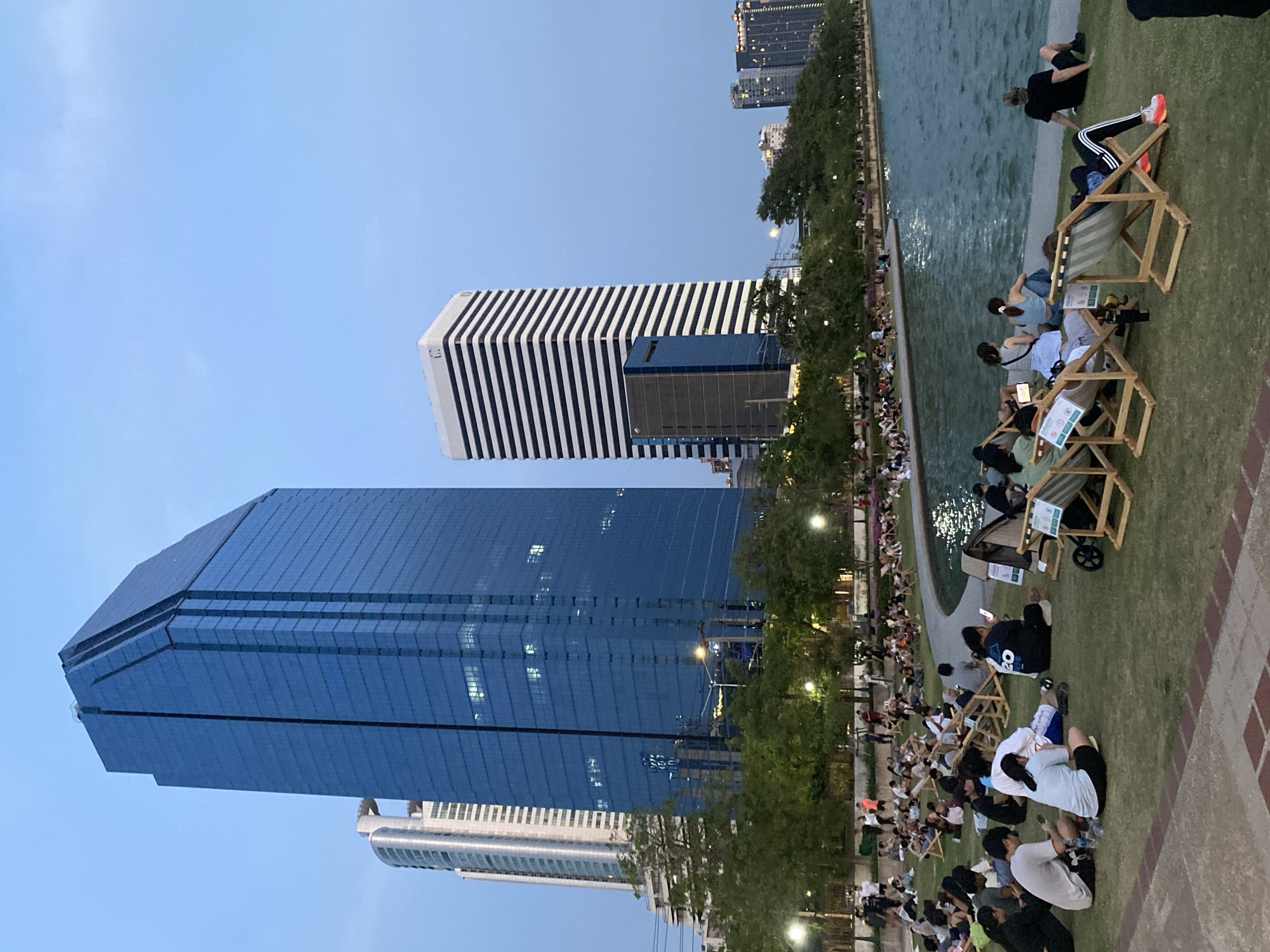 The image shows a park setting in Bangkok, Thailand. Many people are sitting on the grass near a body of water. Some individuals are using small, wooden chairs. Tall buildings are visible in the background, including one with a striking black and white striped design and another covered in blue glass. The sky is a light blue, indicating either early morning or late evening. Trees line the edge of the water. People appear relaxed, looking at their phones and interacting with each other.