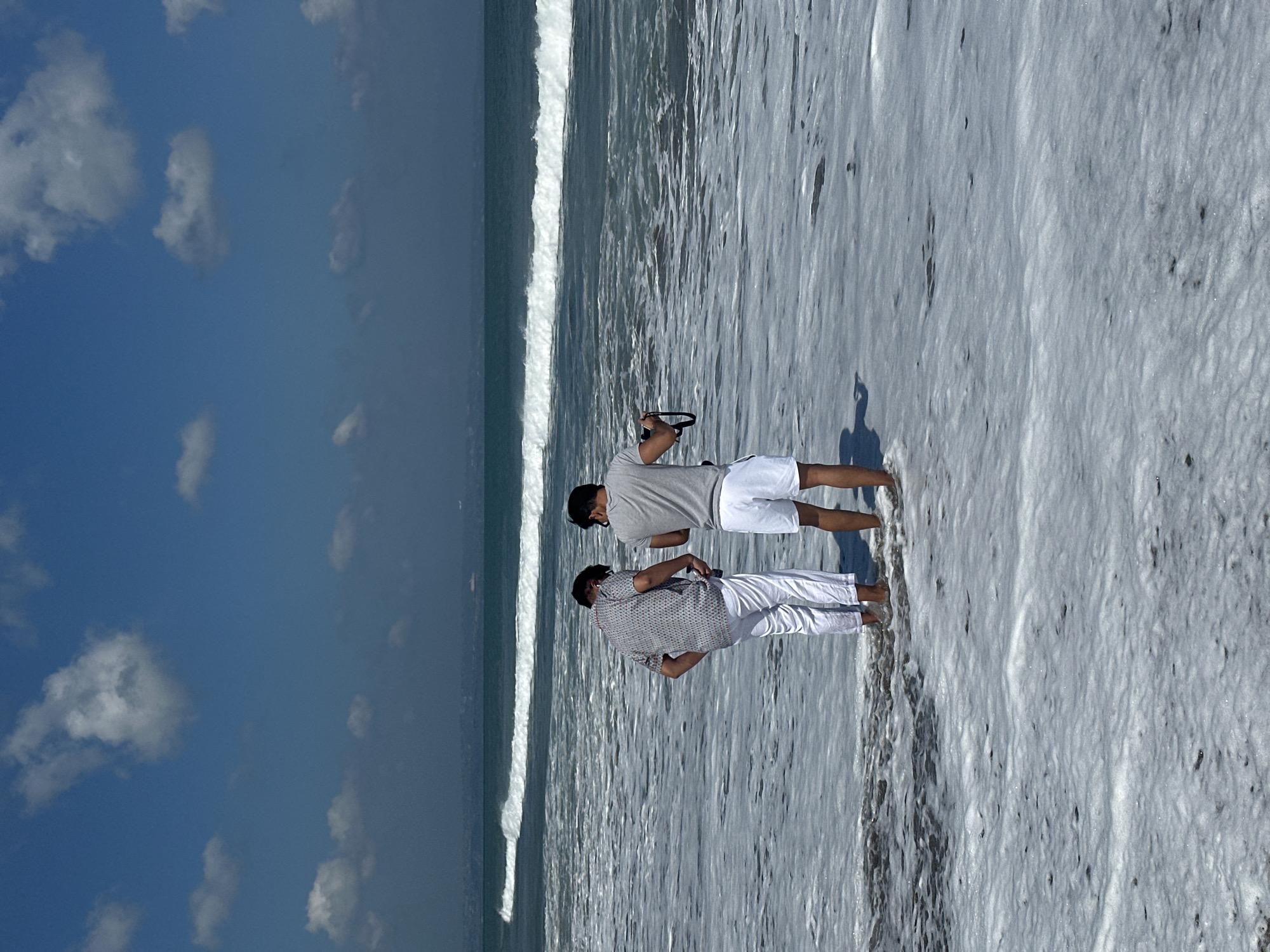 Two individuals are standing in shallow ocean water with foamy waves breaking around their ankles. The person on the left wears a patterned short-sleeved shirt and white pants. The person on the right wears a grey t-shirt and white shorts, and holds a black camera strap. The ocean is a deep teal with a white foamy crest on a breaking wave in the mid-ground. The sky is a clear blue with scattered white clouds. In the far distance, a hazy landmass with a few structures is visible on the horizon. The scene appears to be a beach in North Kuta, Indonesia.