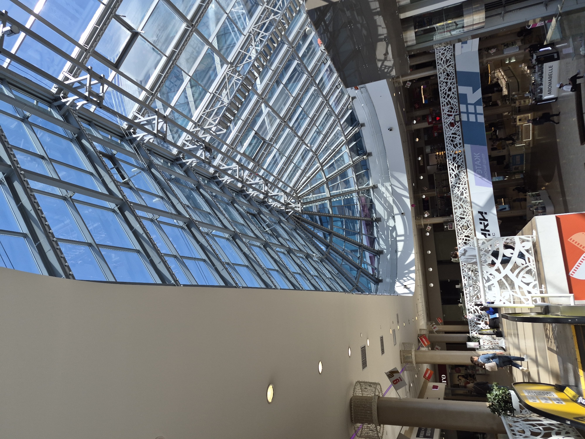The image shows an interior view of a shopping mall in Tobolsk, Russia. The ceiling features a large, arched glass structure with a steel frame, allowing natural light to illuminate the space. Below the glass ceiling is a beige-colored wall with embedded lights. The mall's interior includes pillars decorated with lattice-like patterns, along with signs and banners. People are visible walking and interacting within the mall. Several storefronts and brand advertisements, including one for "Aromabelle," are discernible. Escalators can be seen, and overall, the scene depicts a typical day inside a modern shopping center.