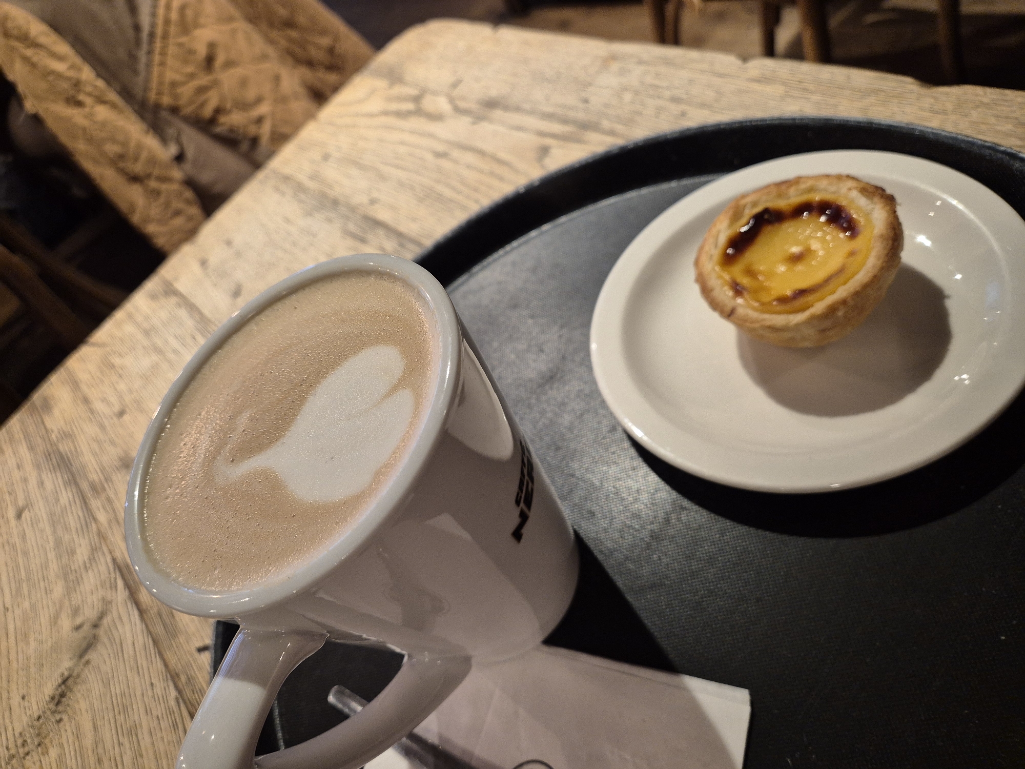 The image shows a close-up of a cappuccino in a white mug and a Portuguese custard tart (Pastel de nata) on a small white plate. Both are resting on a black tray. The tray is on a wooden table, possibly in a café. The location is likely West Suffolk, United Kingdom. There is a chair with a textured fabric covering visible in the background. The cappuccino has latte art in the form of a feather. A white napkin is also visible.