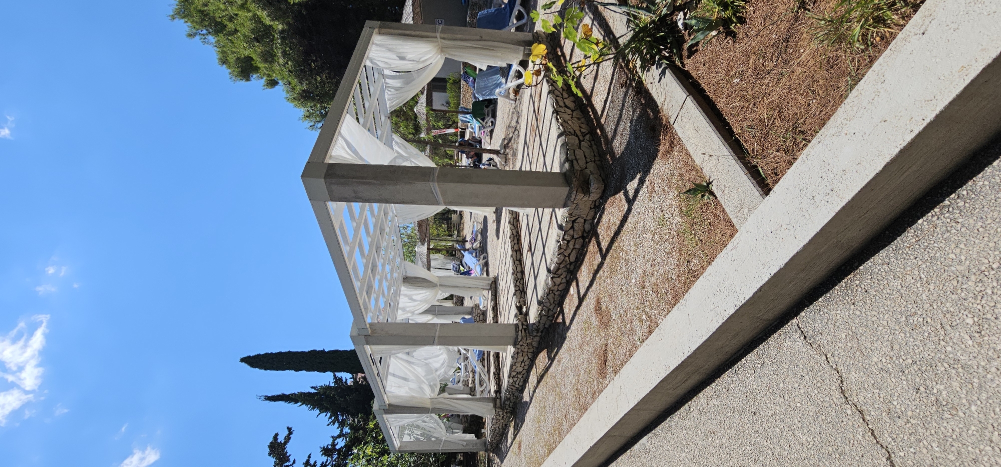 The image shows an outdoor beach or resort area with two covered cabanas. The cabanas are white, rectangular structures with white sheer curtains hanging around them. Each cabana is supported by four concrete pillars and has a slatted roof. The cabanas are situated on raised platforms made of stone.
In the background, several blue lounge chairs are visible, some of which are occupied by people. There are also some thatched-roof umbrellas and some buildings or structures further in the distance.
The ground around the cabanas is covered in gravel and dry grass, with some scattered pine needles. To the left of the cabanas, there is a concrete path or walkway. The area is surrounded by trees, including tall, slender cypress trees.
The sky is clear and blue with a few wispy clouds. The sun is shining, casting shadows from the cabanas onto the ground. The overall scene suggests a sunny day at a coastal resort. The location is identified as being in the city of Grad Supetar, Croatia.