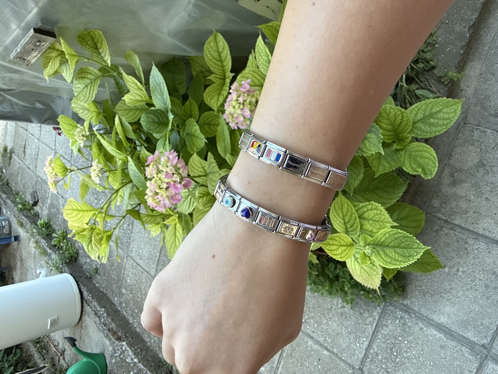 A left forearm wearing two link bracelets is positioned diagonally across the frame. The bracelets are made of silver-colored metal links, each adorned with a charm. The upper bracelet features charms with an evil eye, a lapis lazuli heart, and several other decorative charms. The lower bracelet displays charms depicting flags and other small icons. The forearm is smooth and unblemished. In the background, vibrant green foliage and pale pink hydrangea blossoms are visible. The ground is paved with grey square tiles. To the left, a white cylindrical object and a green watering can are partially in view. The scene suggests an outdoor setting, possibly a garden or patio area. The image does not contain any identifiable landmarks or details to associate it with the city of Tsarevo, Bulgaria.