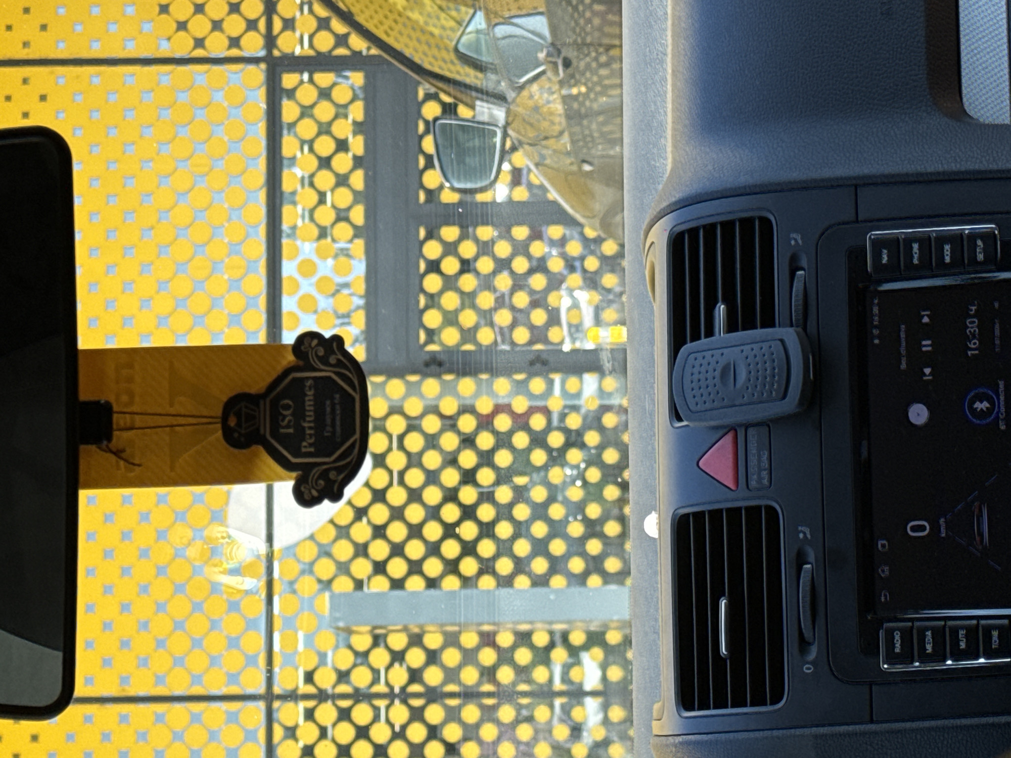 The image shows the interior of a car, looking through the windshield at a yellow, polka-dot patterned building. A car air freshener is hanging from the rearview mirror. The dashboard of the car is visible, including the radio display, air vents, and hazard light button. The time displayed on the radio is 16:30. The exterior scene suggests a modern, possibly commercial or industrial setting, given the geometric pattern of the building. The context provided about Shumen, Bulgaria, indicates this is likely a common sight in that city, but the image itself does not contain any explicit indicators of its location.