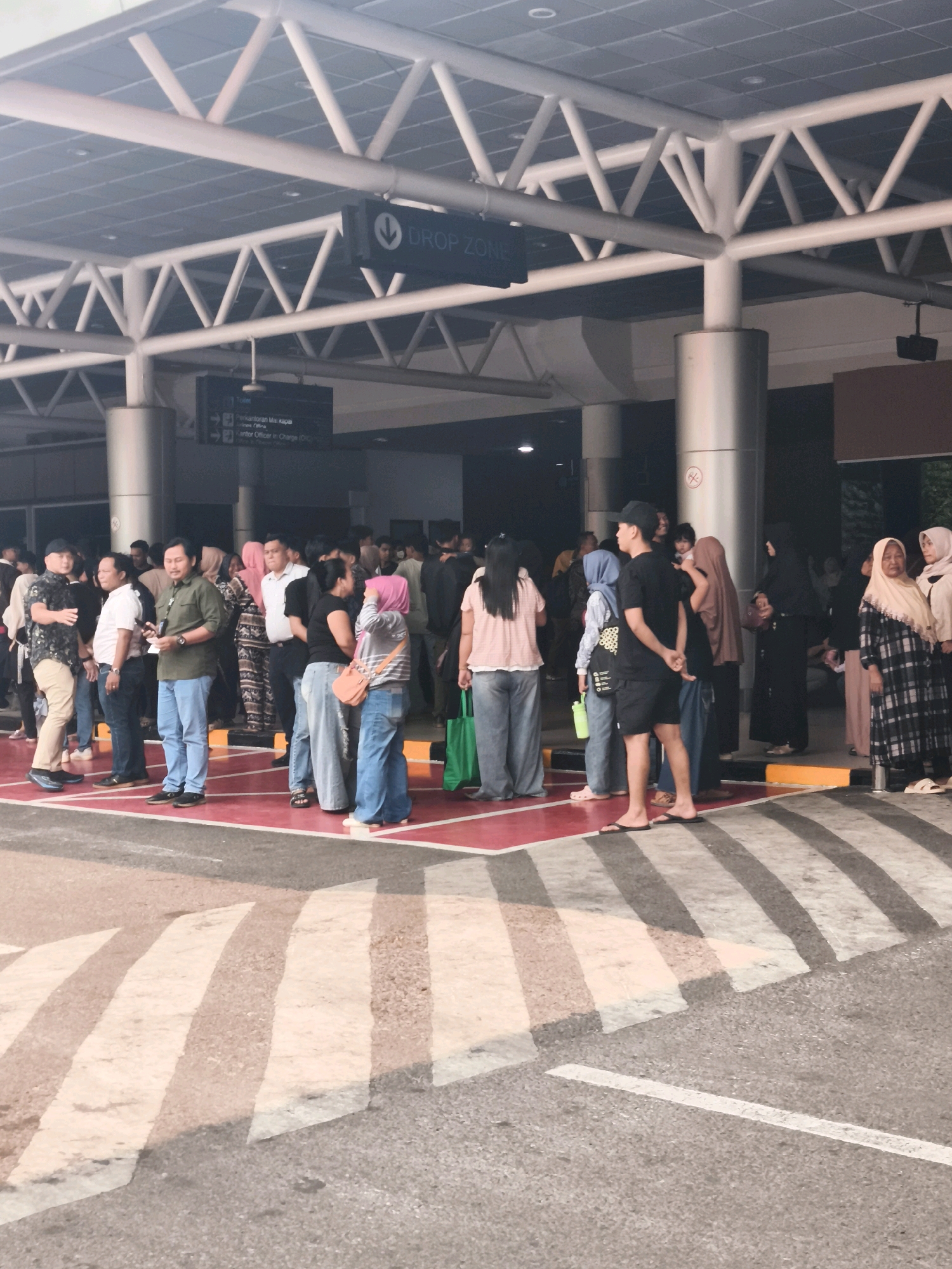 The image depicts an outdoor scene at what appears to be an airport or transportation hub in Palembang, Indonesia. A group of people, both men and women, are standing in a line or gathering under a covered walkway. The walkway is supported by a white metal truss structure, and there are signs visible, including one that reads "DROP ZONE" with an arrow pointing downwards, and another with Indonesian text indicating directions like "Toilet" and "Kantor".
The people are dressed in a variety of casual and formal attire. Some individuals are wearing masks. The ground is a red and gray surface with prominent white pedestrian crossing lines. In the background, a building structure is visible, with some advertising or informational displays. The lighting suggests it might be daytime, possibly overcast or late afternoon, given the overall soft illumination. The environment seems to be a public waiting area, possibly for pick-up or drop-off services.