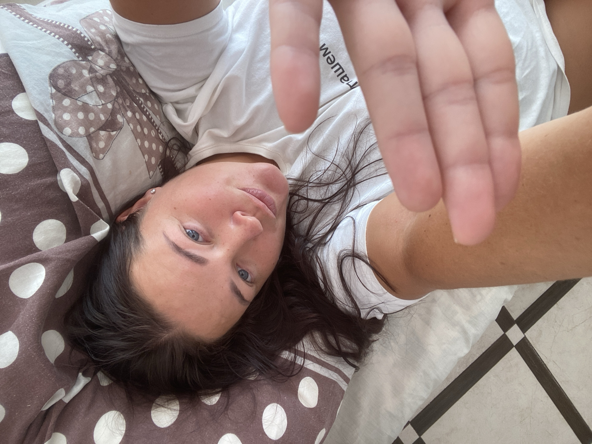 A woman is lying on her back in a bed. Her head is turned to the side and her eyes are open. She is wearing a white t-shirt and her hand is held up in front of her face, obscuring part of the image. The pillow she is resting on has a brown and white polka dot pattern. The bed is on a tiled floor. It's difficult to ascertain the specific location within Sochi, Russia without further context, but the scene suggests an indoor domestic setting.