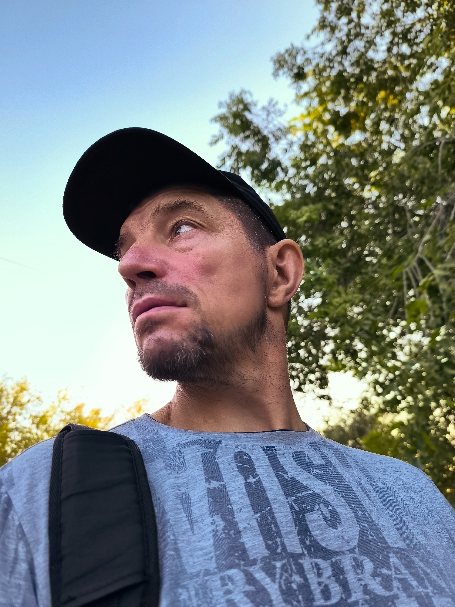 A low-angle, medium shot captures a man from the chest up, looking to his left against a bright, hazy sky and out-of-focus green foliage. The man wears a black baseball cap and a light blue t-shirt with a dark graphic print. He has short, dark stubble on his chin and a fair complexion. A portion of a black backpack strap is visible on his left shoulder. The background suggests an outdoor setting with trees. The scene is consistent with a candid outdoor photograph, likely taken during daylight hours.