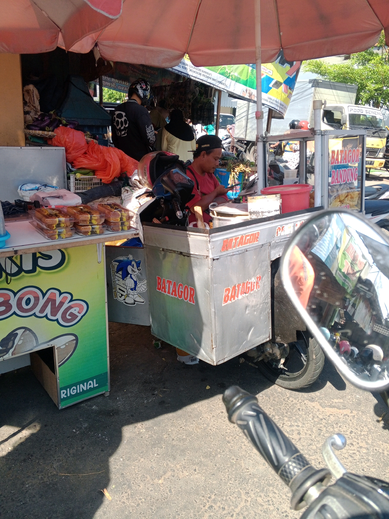 An outdoor food stall, likely a "Batagor" stand, is set up on a paved area, possibly a roadside or market in Wonogiri, Indonesia. The stall features a red and white striped umbrella providing shade over the workspace. Several containers of food, packaged in clear plastic, are displayed on a white counter. Behind the counter, a person wearing a red shirt and a black cap is seen preparing food. Various items, including baskets of produce like eggplants and oranges, and red plastic bags, are stored around the stall. A silver metal cart with "BATAGOR BANDUNG" written on it in red letters is positioned in front of the stall. The scene is framed by the reflection in a motorcycle's side mirror, which shows other stalls, people, and vehicles, hinting at a bustling environment. The ground is asphalt, with some scattered debris.