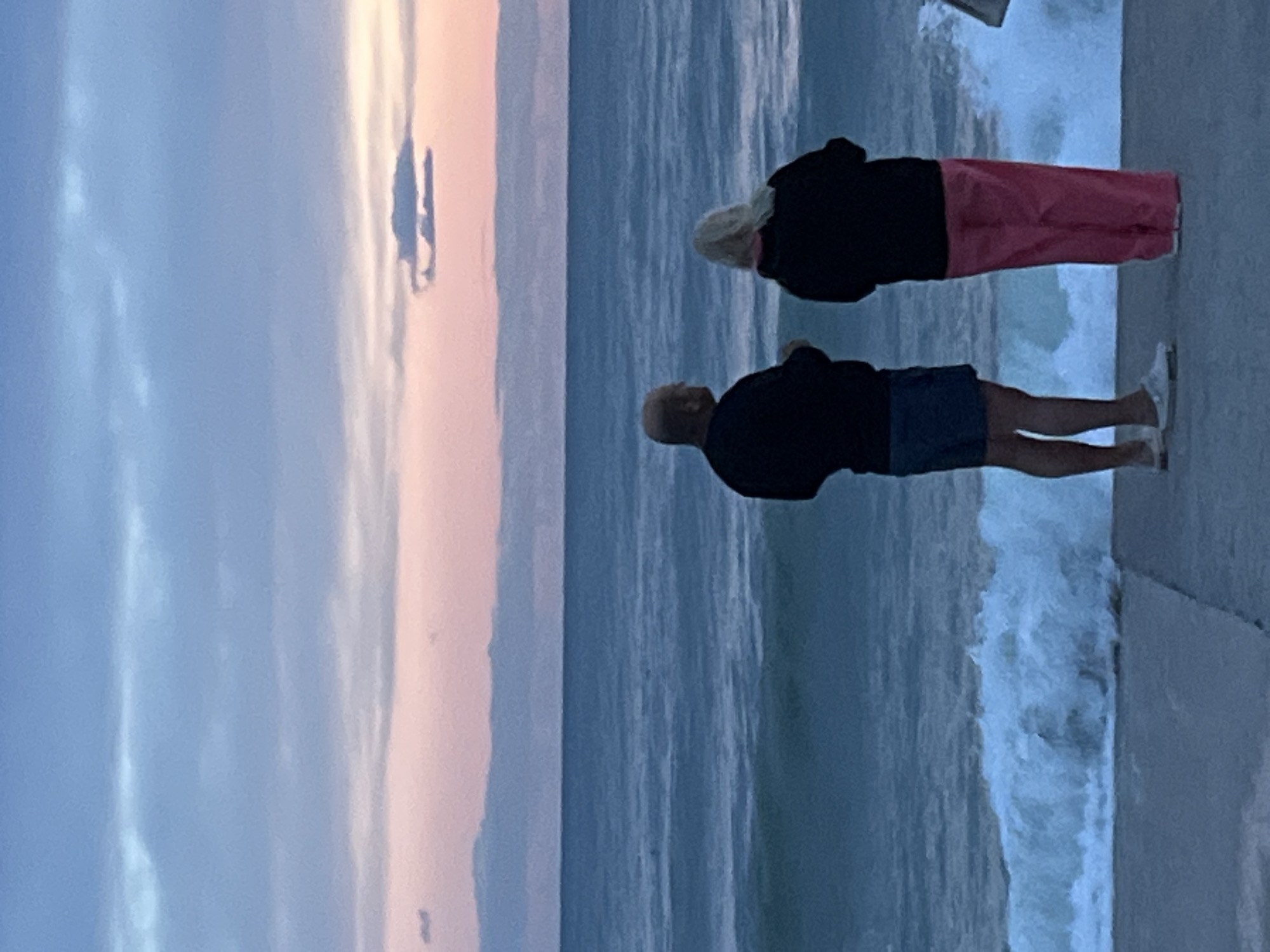 Two people, an adult male and an adult female, stand on a concrete pier or promenade at the edge of the ocean. The man wears a dark shirt and blue shorts. The woman wears a dark shirt and pink pants. Both are facing the ocean with their backs to the camera. The sea is turbulent, with waves crashing against the pier. The sky is a mix of blues and pinks, suggesting either dawn or dusk. A figure appears to be in the distance above the water. The location is Alanya, Turkey.