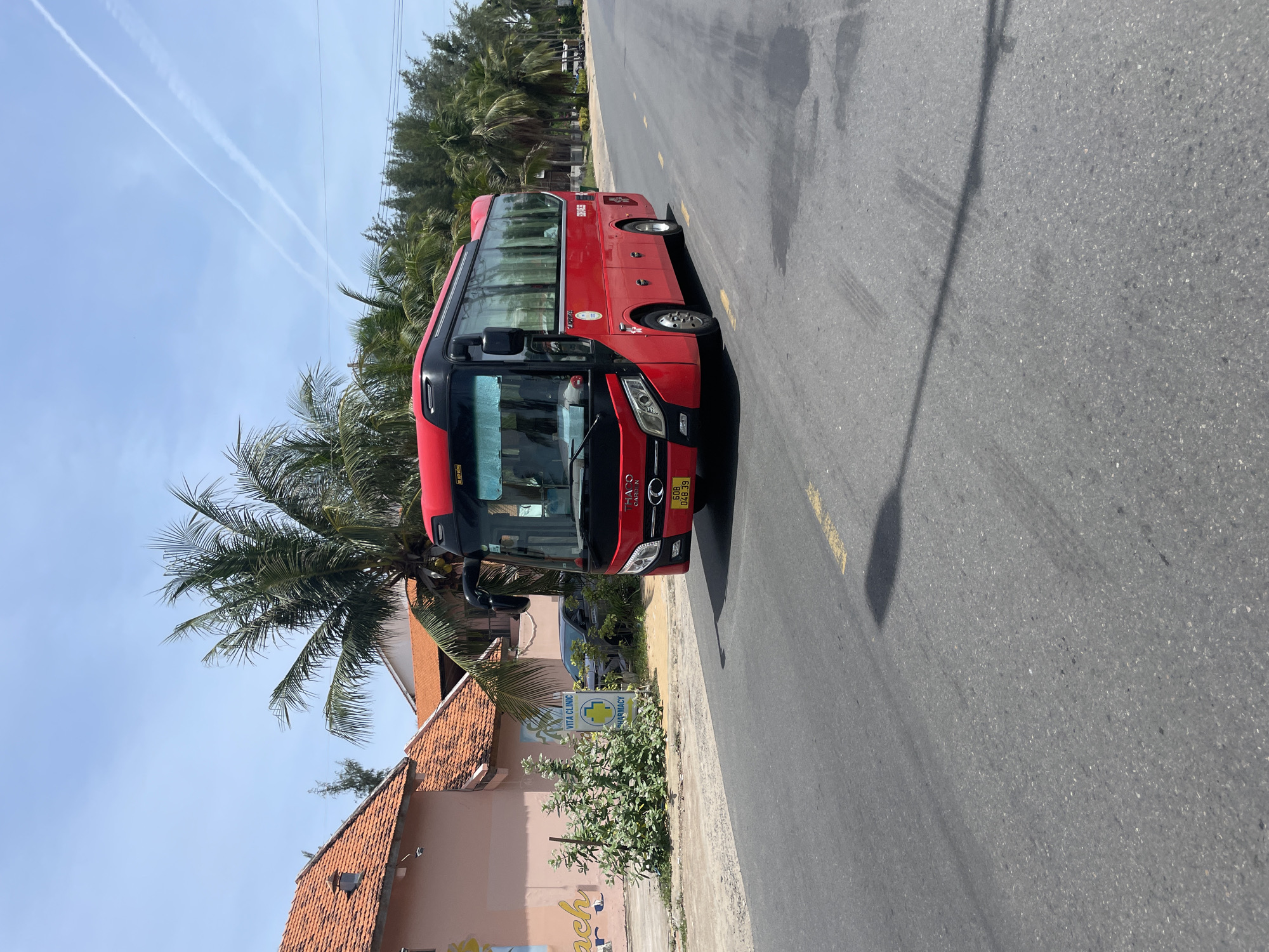 A bright red passenger bus is traveling on a two-lane asphalt road in Phan Thiet Ward, Vietnam. The bus has tinted windows and is labeled with Vietnamese characters. On the left side of the road, there are palm trees, a light pink building with an orange tiled roof, and a sign that reads "VITA CLINIC PHARMACY." In the background, more palm trees and foliage line the side of the road. The sky is blue with some faint white contrails from an aircraft. The road has yellow lane markings and some dark patches of wetness or oil stains. The bus is captured in motion, with its tires on the asphalt. The overall scene depicts a sunny day in a coastal or tropical environment.