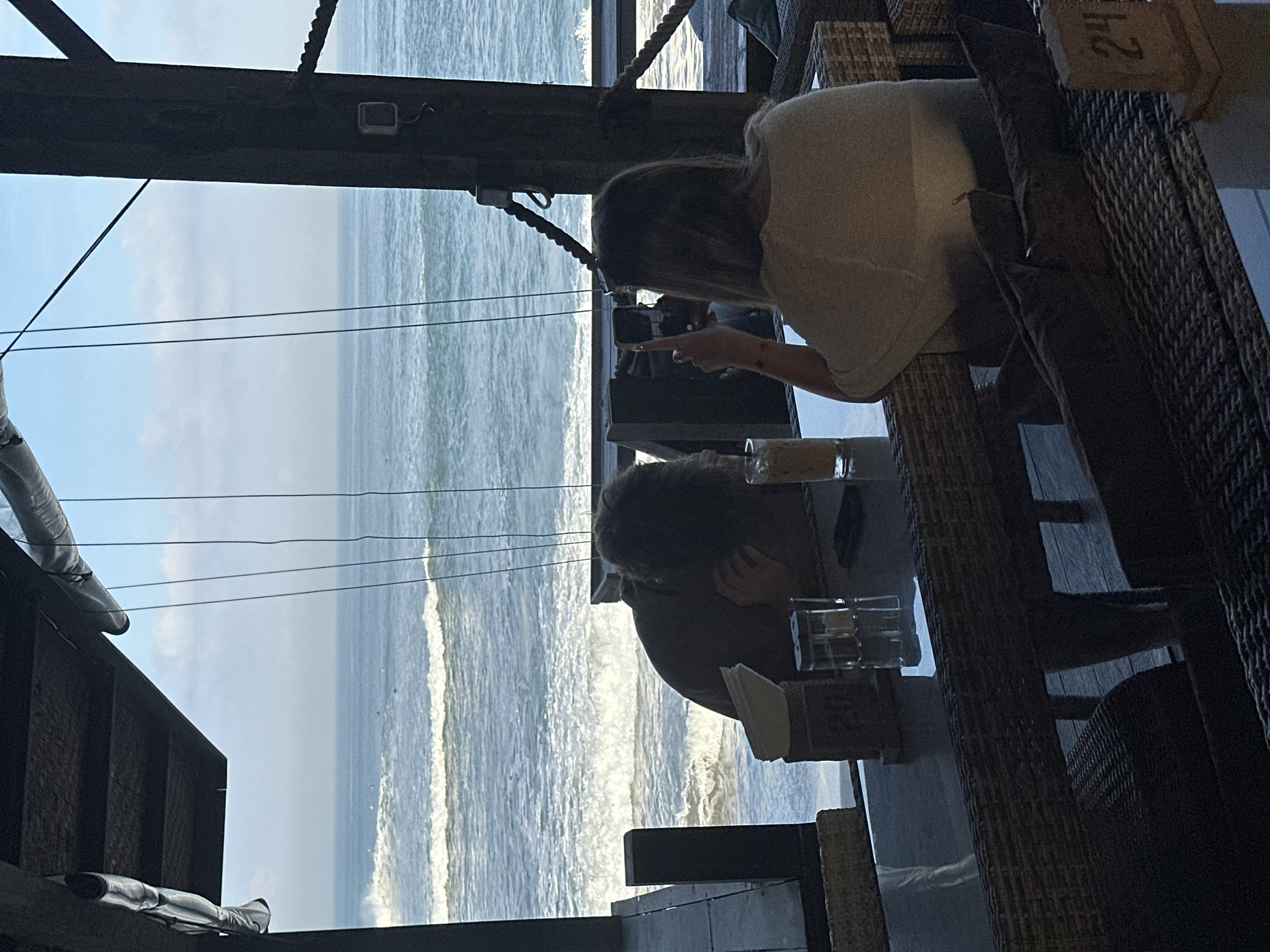 This is a vertical, eye-level shot of two people seated at a table at a restaurant overlooking the ocean in North Kuta, Indonesia. The scene is framed by a dark, weathered wooden overhang and a thick wooden pillar. The ocean is visible beyond the table, with waves breaking and several surfers in the distance. The sky is a light blue with some scattered clouds.
In the foreground, a glass table is set with a napkin dispenser, salt and pepper shakers, a phone, and two drinks. A person with dark, curly hair is slumped over the table, facing away from the camera and towards the ocean. Their face is obscured by their hair. To the right, a person with blonde hair, wearing a light-colored top, is holding up a smartphone and appears to be taking a picture or video, with their index finger extended. They are facing away from the camera, also towards the ocean. The seating area features woven rattan chairs and benches. A small wooden sign with the number "24" is visible on the right side of the frame.