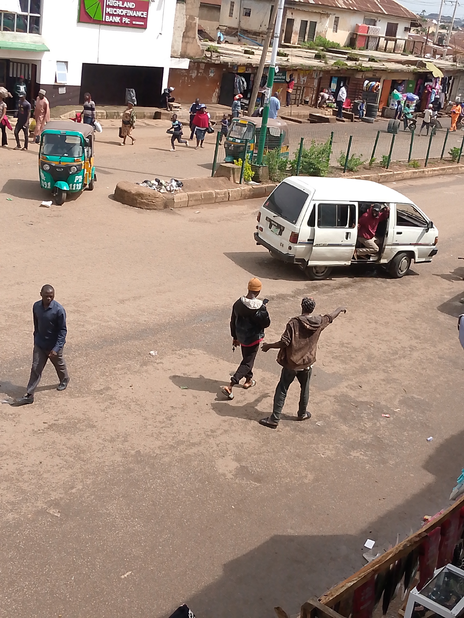 This image captures a street scene in Bukuru, Nigeria, likely near a commercial area given the buildings with storefronts and signage. In the foreground, a man in a dark blue shirt and grey pants walks from left to right, casting a shadow. Behind him, two men walk towards the right. One, wearing a brown hooded jacket and dark pants, points to the right. The other, in a black jacket and tan beanie, holds a phone. To the left, a green and blue auto-rickshaw is parked. Further in the background, a white van with its side door open is parked, and a person in a red garment is visible inside. Several other individuals are seen walking and standing in the midground, some near market stalls or shops. The ground is paved with asphalt. The environment appears to be a bustling urban setting with buildings of varying states of upkeep.