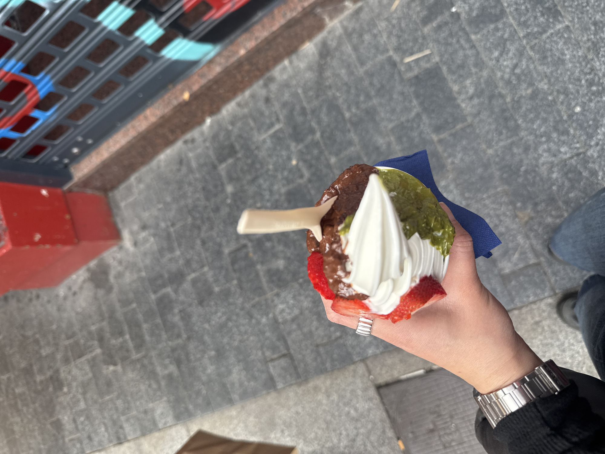 The image shows a person holding a dessert in their hand, likely an ice cream or yogurt parfait, consisting of layers of chocolate, strawberries, white soft serve, and green topping. They are using a wooden spoon to eat it. The person wears a silver wristwatch on their left wrist. They are standing on a paved street, and a portion of a storefront with a red and blue design is visible in the background. While it's difficult to pinpoint the exact location in Madrid with just this image, the urban paving style is typical of Madrid's streets.