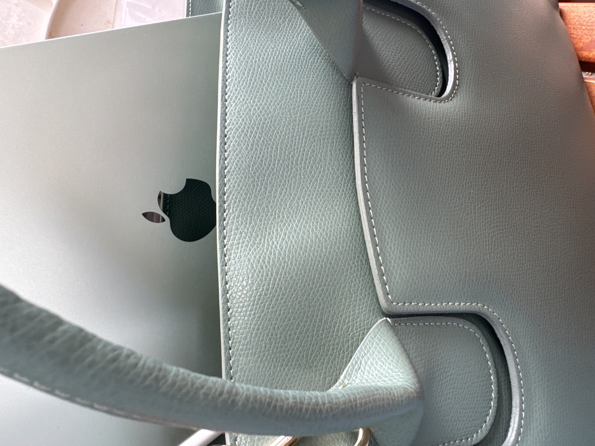 The image shows a silver Apple laptop with a black Apple logo partially inside a light green leather tote bag. The bag has a handle and stitched details. The setting is unclear, with only hints of other objects visible in the background. The location of the image is not discernible based on the information provided.