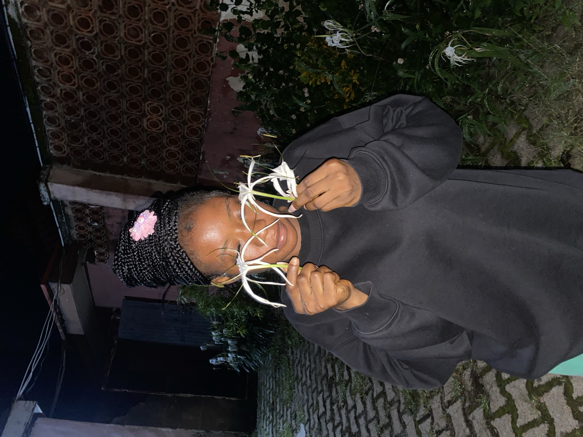 The image shows a young Black woman with braided hair, adorned with a pink flower, holding two white spider lilies to her face. She is wearing a black sweatshirt and appears to be outdoors at night. The background includes a brick wall with a patterned design, vegetation, and a paved walkway. The overall impression is one of a relaxed and artistic pose in a dimly lit environment.