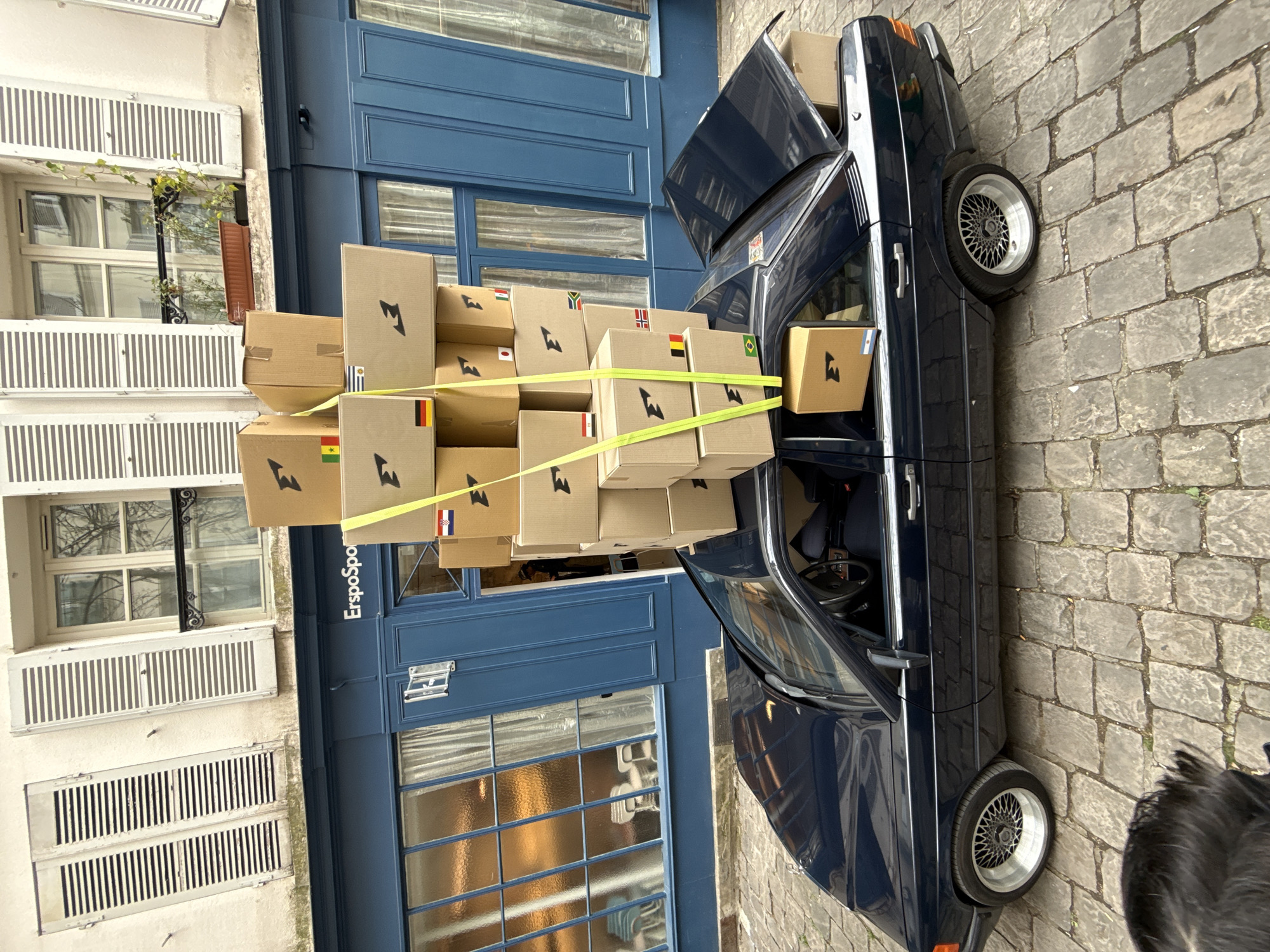 The image shows a blue car loaded with cardboard boxes, secured with yellow straps, parked on a cobblestone street in Paris, France. The car's trunk is open, revealing additional boxes. The boxes are marked with a logo resembling a lightning bolt and adorned with small flags from various countries, including Brazil, Germany, Greece, Japan, South Africa, and others. The vehicle is parked next to a building with white shutters and a blue storefront, on which a sign appears to read "ErspoSpot". The architecture suggests a traditional Parisian street. Part of a person/animal with black fur is visible on the lower right corner.