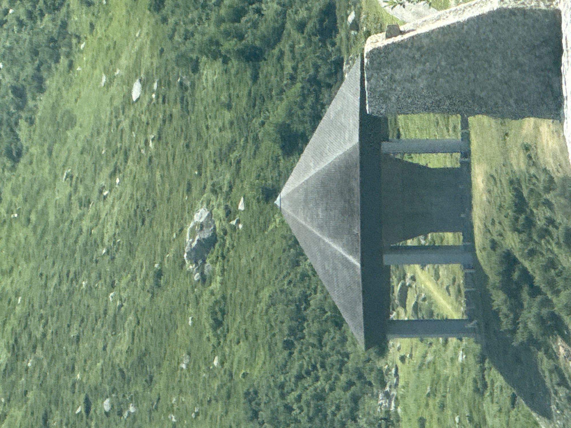 The image depicts a hexagonal, grey, multi-level structure with a dark roof, supported by four cylindrical pillars. The structure is situated on a grassy hillside, with a path winding behind it. To the right of the structure, a large, rough-hewn stone monument stands. The background is a lush, green, mountainous landscape with scattered rocks and trees. The scene appears to be a public space or a monument, likely in a rural or mountainous area. This specific structure is the Walter Gropius Memorial, a tribute to the renowned architect, located in the Swiss municipality of Hospental, within the canton of Uri. The memorial was designed by Gropius himself and was unveiled in 1967.