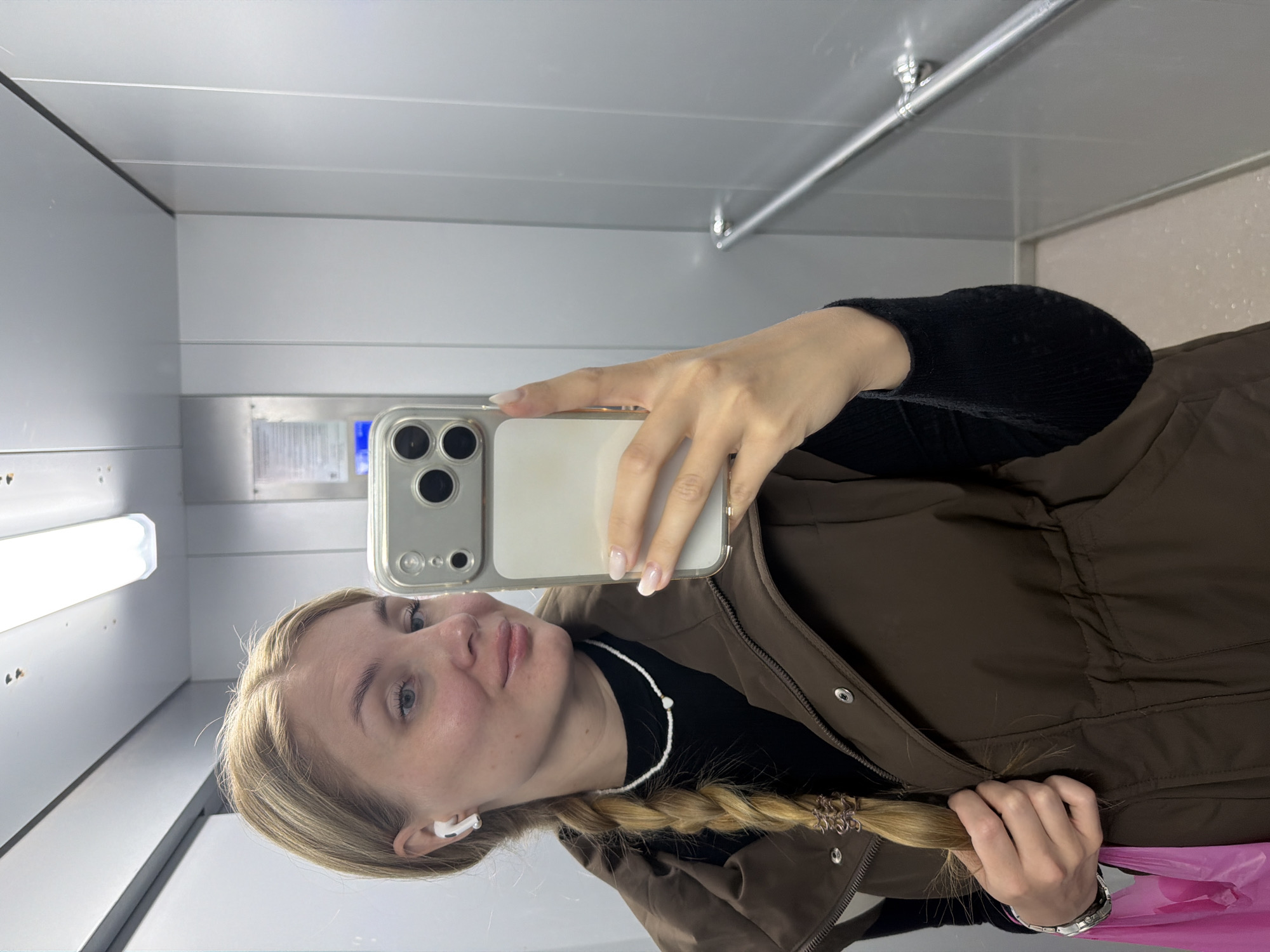 A young woman is taking a selfie in what appears to be an elevator. She is holding a cell phone in her right hand, the phone is angled toward her face. She is wearing a brown jacket over a black top and a white beaded necklace. Her blonde hair is styled in a braid. The elevator interior consists of gray metal paneling, with a long light fixture and a metal bar along one wall. An elevator call button panel is partially visible in the background. Based on previous information, the location is presumed to be in the city of Chaikovsky, Russia.