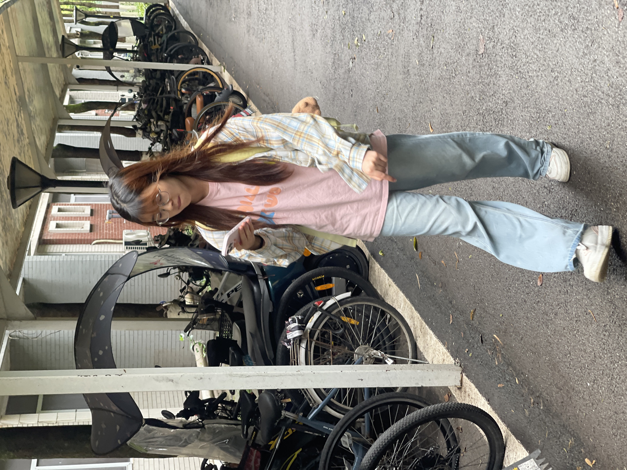 A young East Asian woman walks with a phone in her hand, looking down at it. She is wearing a pink t-shirt with orange text, a light-colored plaid shirt, light blue wide-leg jeans, and white sneakers. She has long, dark brown hair and is wearing glasses. She has a yellow backpack over her left shoulder and a small, brown, plush toy attached to her right. The woman is walking on a paved path under a covered overhang, with many bicycles parked in a row to her left. The environment appears to be a bike parking area.