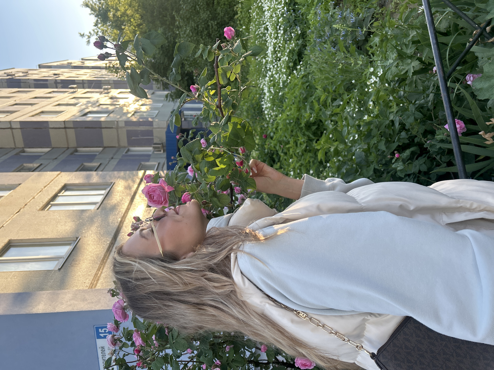 The image, taken during the golden hour of a sunny day, depicts a woman interacting with a rose bush in an urban environment in Tolyatti, Russia. The woman, shown in profile from the waist up, has long blonde hair and is wearing sunglasses. She is dressed in a light-colored hoodie or sweatshirt layered under a puffy vest or jacket, and a gold chain strap with an "MK" logo from a handbag is visible. Her head is tilted towards a pink rose, suggesting she is smelling or admiring it.
In the background, a multi-story residential building with a light-colored facade and distinctive purple-blue horizontal striping is visible, with sunlight reflecting off its windows. To the right of the woman, a dense growth of green foliage, including more pink roses and small white and blue flowers, forms a natural backdrop.
At the bottom left, a blue street sign is partially visible, bearing white text that reads "строителей" (stroitel'ev, meaning "builders") and the number "15," indicating a street address. The warm, low sunlight highlights the woman's hair and parts of the building, creating a pleasant, contemplative atmosphere.