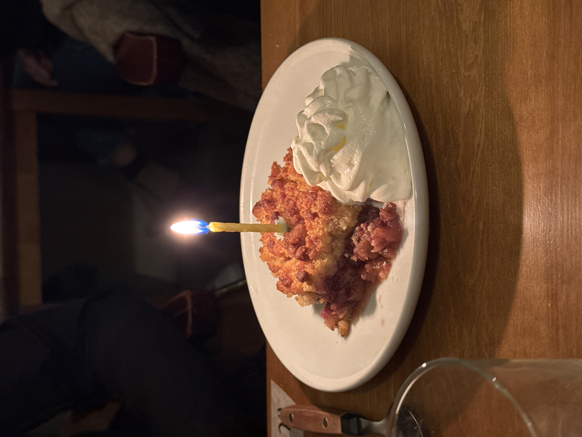 The image displays an indoor, low-light scene centered on a dessert placed on a light-brown wooden tabletop. A white, round ceramic plate holds a slice of fruit crumble featuring a golden-brown, textured topping and a base of reddish-purple stewed fruit. A single, lit yellow candle, emitting a flame with a blue base and orange tip, is embedded vertically into the crumble. Adjacent to the crumble, on the right side of the plate, is a dollop of white whipped cream.
In the lower left foreground, a partial view of a clear glass tumbler is visible, along with the handle and blade of a metal utensil, possibly a serving knife or spatula, with a dark wooden handle. The background is dark and significantly out of focus, showing blurred figures of at least two individuals seated. One figure on the left appears to be wearing dark clothing, while another on the right is in lighter, textured attire. Indistinct reddish-brown objects, possibly parts of chairs or furnishings, are also faintly discernible in the background. No specific visual elements within the image provide identifiable information regarding its precise location in Paris, France.