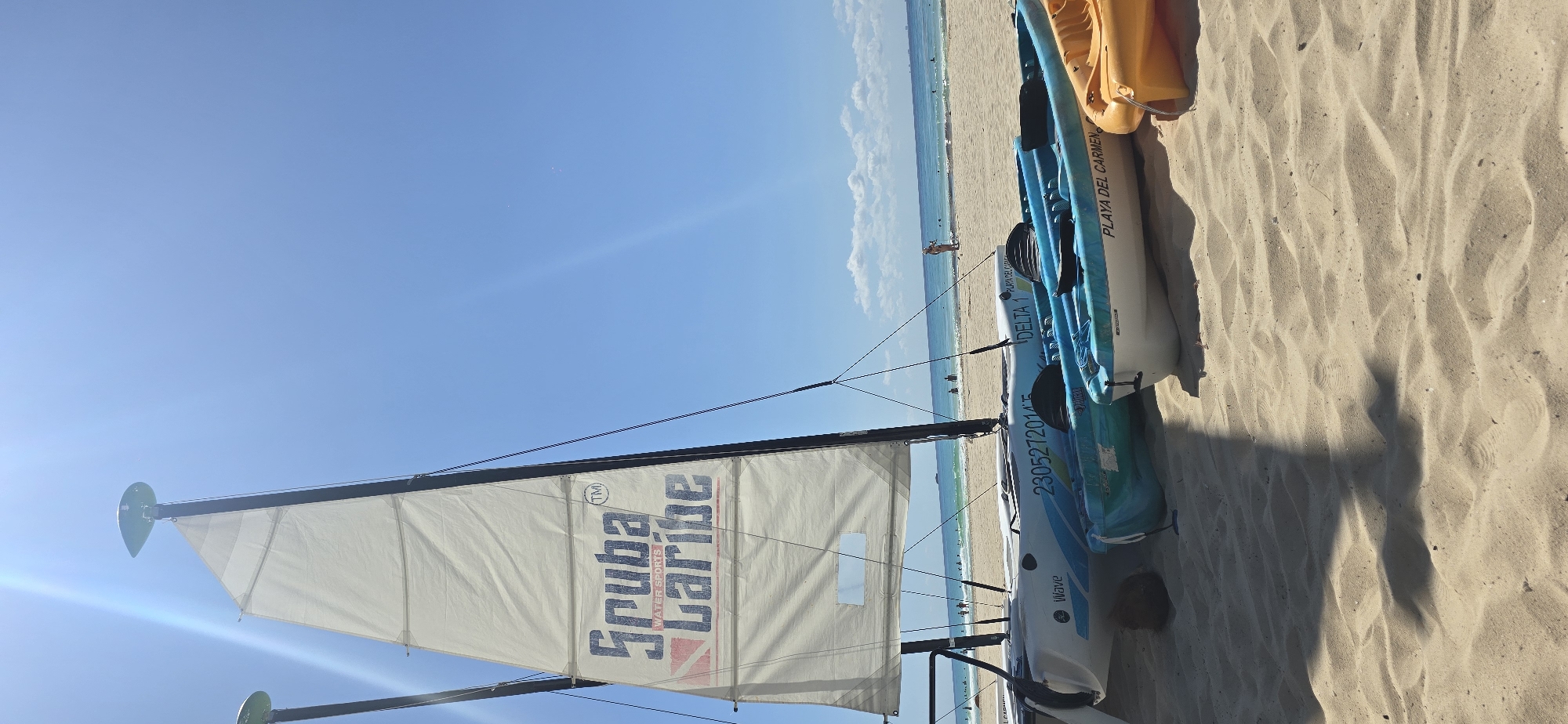 The image captures a beach scene with a sailboat and kayaks resting on the sand in Playa del Carmen, Mexico. The beach is sandy, with visible patterns created by wind and water action. Two kayaks are stacked on top of each other. The top kayak is yellow, and the lower one is white and light blue and has the text "PLAYA DEL CARMEN" and "DELTA 1". In the background is a sailboat with a white sail that reads "Scuba Caribe" with an apparent TM symbol nearby, indicating a water sports company. In the background, there is a view of the ocean and the coastline, where people are visible on the shoreline. The sky is clear and blue.