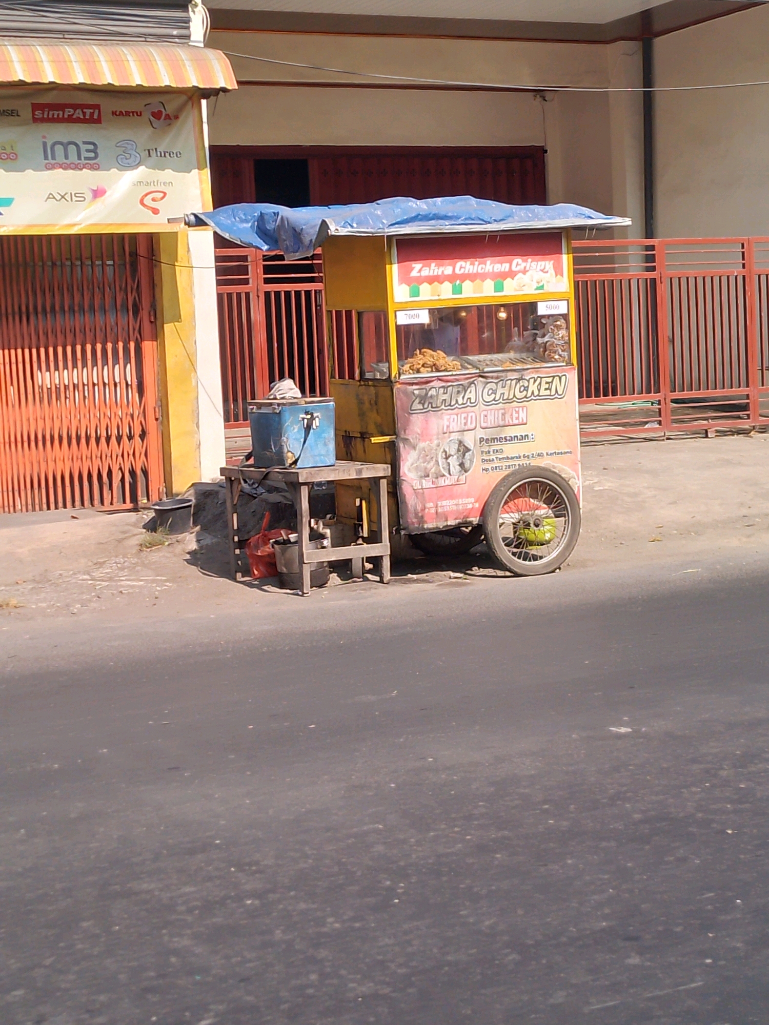 An eye-level, medium shot captures a food cart parked on the side of a road in Kertosono, Indonesia. The cart, branded "Zahra Chicken Crispy Fried Chicken," is adorned with colorful banners and displays a variety of fried chicken. The background reveals a building with red metal gates and a yellow facade, suggesting a commercial area. Sunlight casts shadows on the asphalt road and the pavement, indicating a daytime scene. The cart is equipped with a wheel, likely for mobility, and a small table with containers. The overall atmosphere is that of a typical street food vendor operation in a bustling Indonesian city.