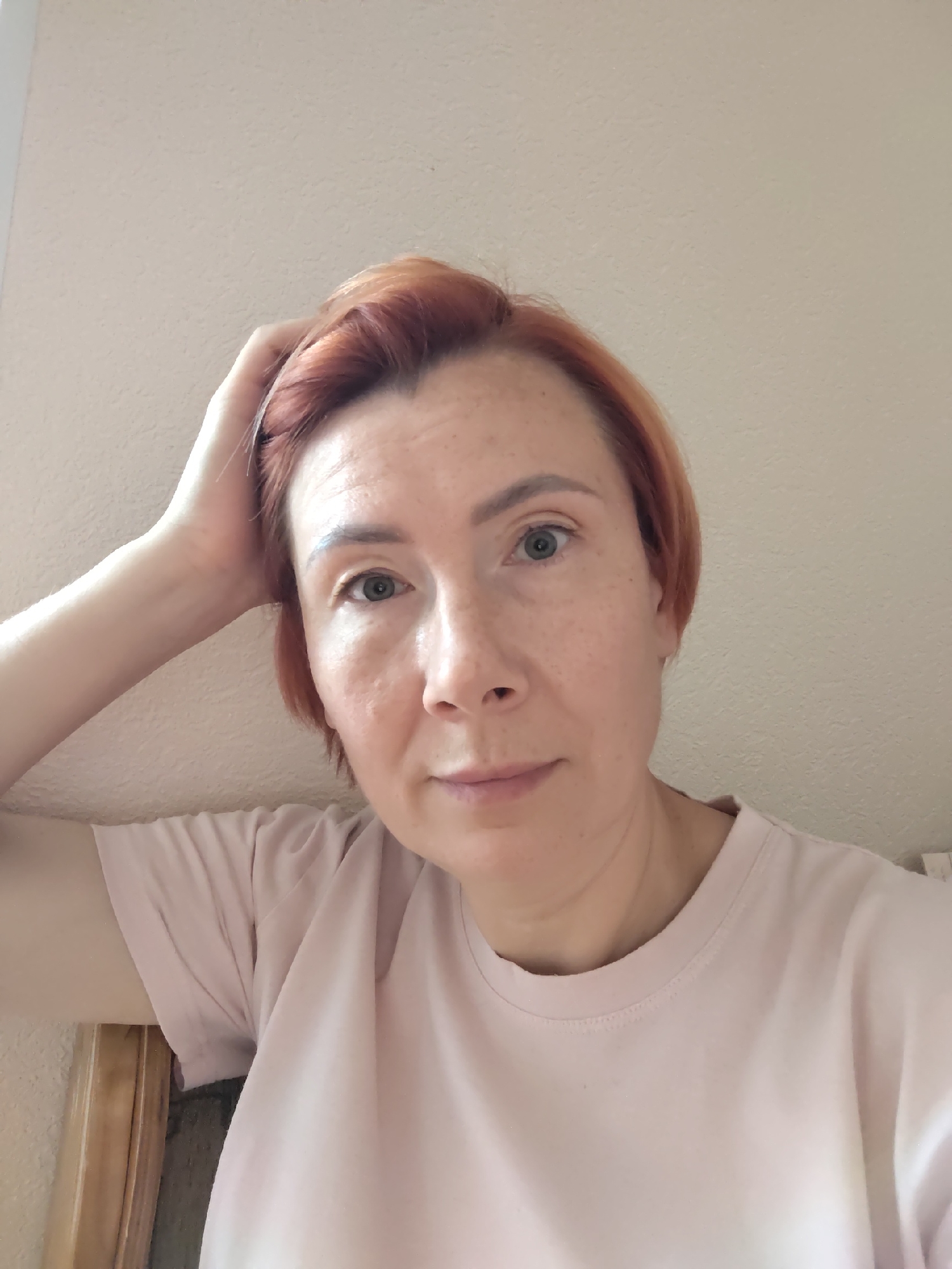 A woman with short, reddish-brown hair is looking into the camera, resting her right hand on her head. She is wearing a light pink t-shirt. Her skin has freckles and is slightly flushed. The background appears to be a textured, light-colored wall. The image is a close-up selfie, with the woman's face and upper body filling most of the frame. There are no specific objects or landmarks visible that would indicate a precise location within Konakovo, Russia.