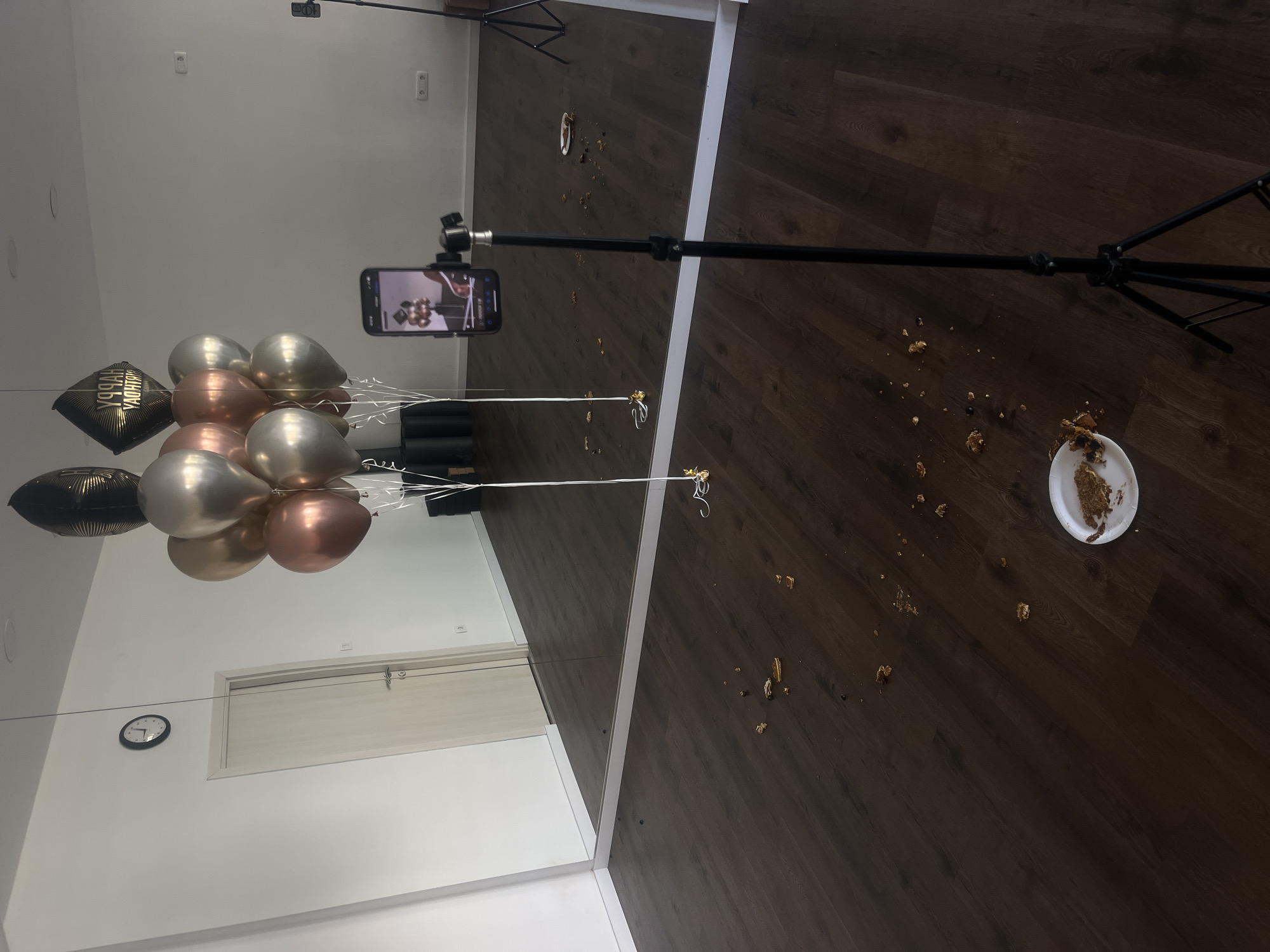 Indoor scene, likely a studio or event space in Budva, Montenegro, judging by the décor and likely rental nature of the location. The primary feature is a large mirror encompassing a significant portion of a wall. Reflected within the mirror are several metallic balloons (silver, gold, rose gold, and black with gold lettering that appears to say "Happy Birthday") and a wall clock.
A smartphone is mounted on a tripod, positioned to capture the scene, presumably recording a video of the balloons.
Notably, the dark wood flooring has cake debris scattered around a white plate with a portion of cake remaining on it. The cake’s crumbs and remnants suggest a recent incident, implying the cake was dropped or partially consumed. The floor appears clean apart from the cake and crumbs.
The overall impression is of a potentially celebratory event cut short by an accident involving the cake.