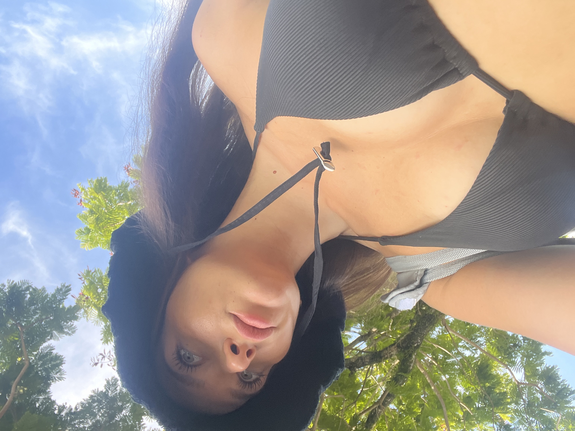 A woman wearing a black bikini and a black hood is the main subject of this low-angle photograph. The background features green trees and a blue sky with some clouds, suggesting an outdoor location. Given the prompt's mention of Choeng Thale, Thailand, the location is likely a park or beach area within that city. The woman is looking down at the camera, creating a selfie-style composition.