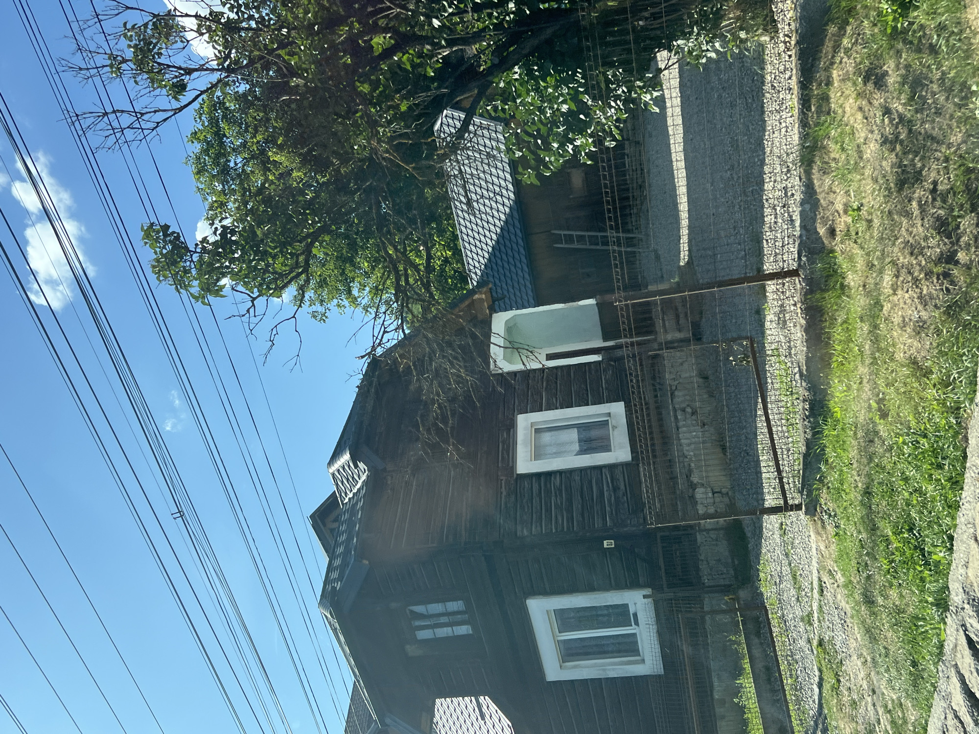 The image depicts a wooden house with a dark-stained exterior and a dark, tiled roof, situated in the city of Prisaca Dornei, Romania. The house has several windows, some with white frames. A smaller, adjacent building with a similar dark, tiled roof is also visible. A metal fence runs along the front of the property, partially obscuring the yard which is covered in gravel and grass. Power lines are strung across the bright blue sky above. Lush green trees frame the right side of the image. The overall scene suggests a residential property in a rural or semi-rural setting.