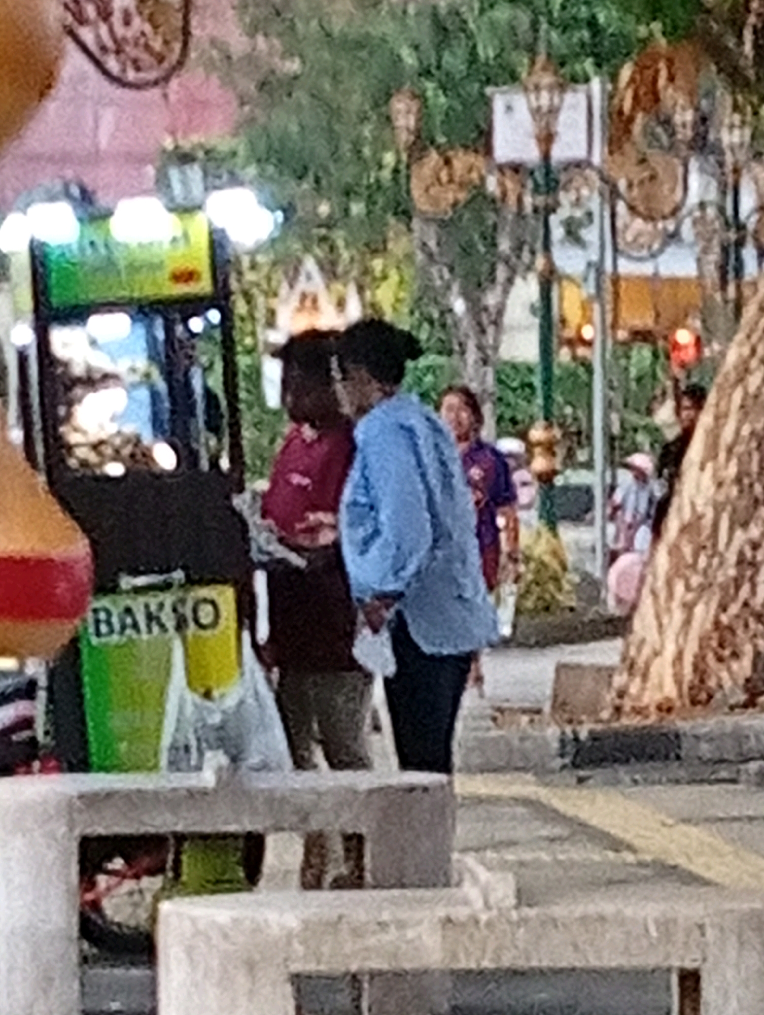 The image captures a street scene in the city of Malang, Indonesia. In the foreground, a white stone structure with multiple compartments is visible. To the left, a food cart with green and yellow containers, one labeled "BAKSO," is present. Two individuals are standing near the cart, one wearing a maroon shirt and the other a blue denim-like shirt. Behind them, other people and a tree with rough bark are visible. Traffic lights are illuminated in the background, suggesting it is nighttime or dusk. The overall impression is that of a bustling street food area.