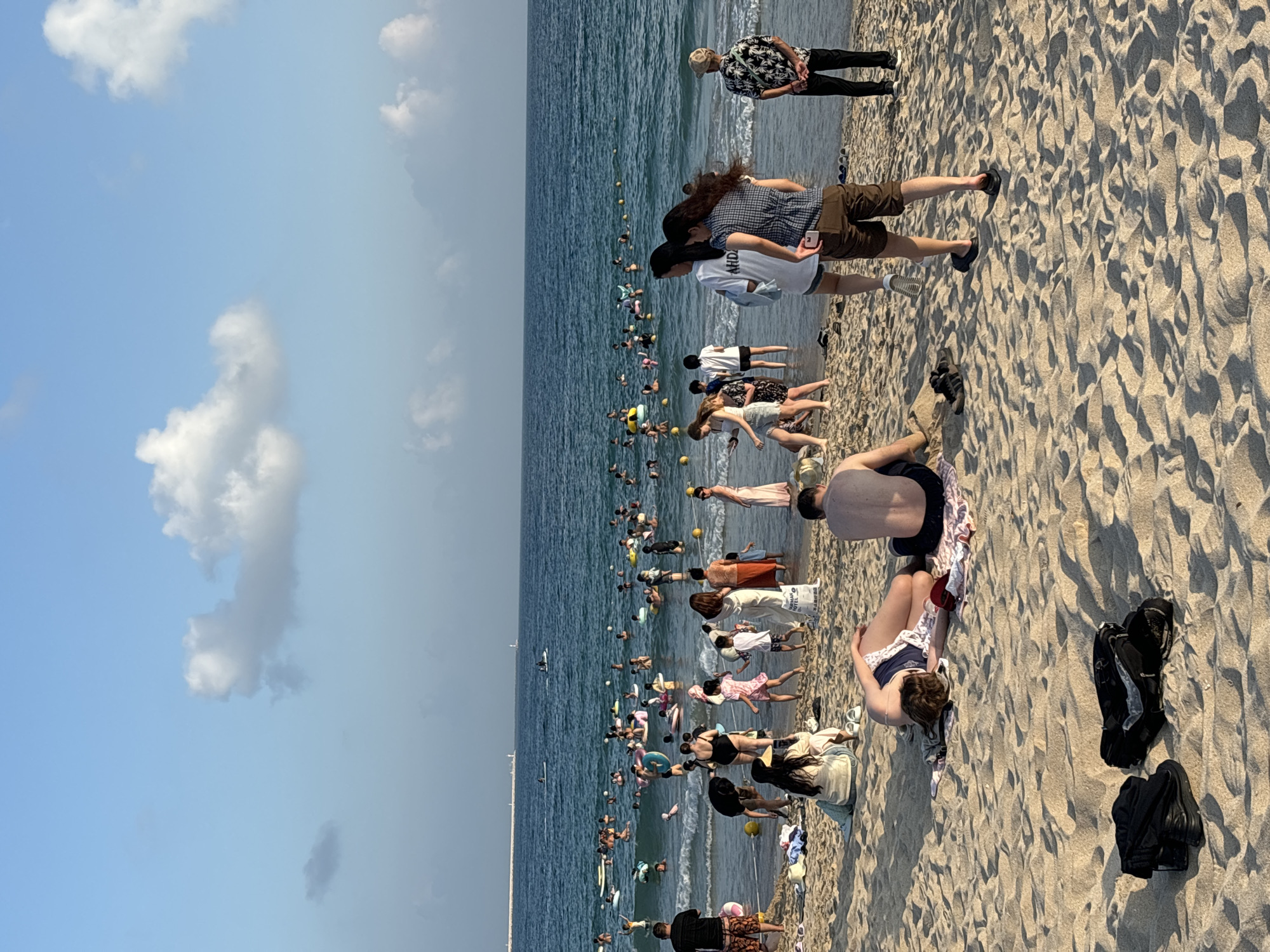 The image depicts a crowded beach scene in the city of Jiyang District, China. The sandy beach is populated by numerous individuals engaged in various activities. In the foreground, two people are lying on towels, one with their head down and the other with their back to the camera. Two pairs of black footwear are placed on the sand in front of them. Further back, a group of people are standing near the water's edge, some are walking into the sea, and others are already in the water, many with inflatable rings. The ocean is blue with some small waves and yellow buoys are visible in the water. The sky is a clear blue with scattered white clouds. In the distance, a long pier or breakwater extends into the sea with a lighthouse structure at its end. Several boats and individuals on paddleboards can be seen on the water further out. The overall impression is a lively summer day at the beach.