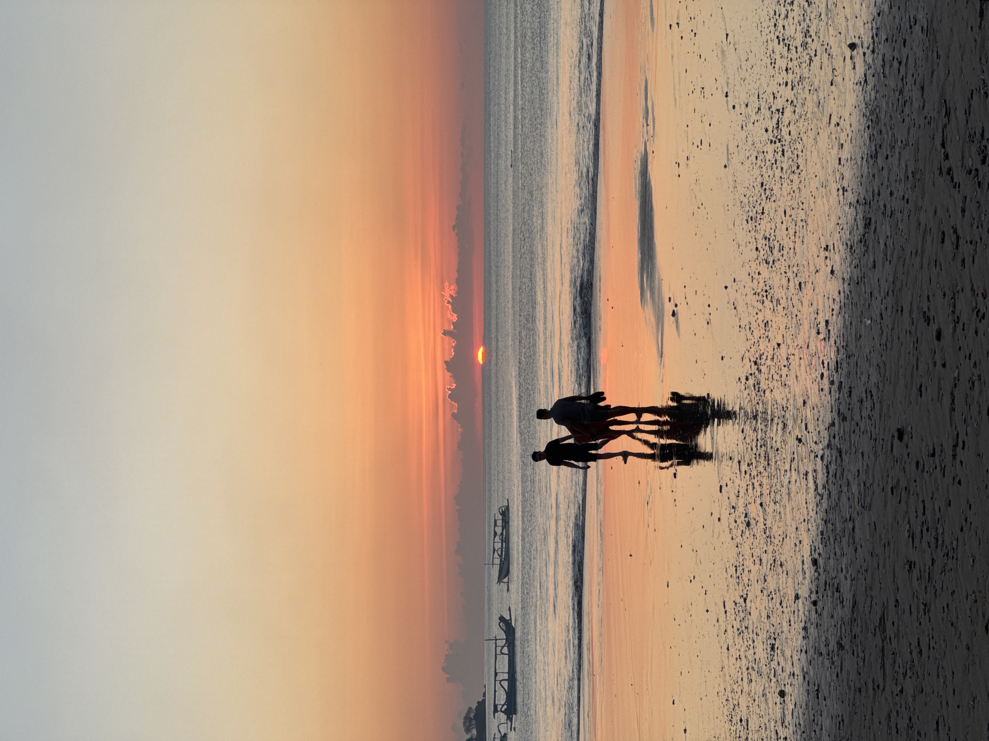 At sunset in Kuta Selatan, Indonesia, two individuals are silhouetted against the horizon as they walk along the wet sand of a beach. The sky is painted with gradients of orange and pale blue, with the sun appearing as a small, bright orb just above the clouds. The calm ocean reflects the warm colors of the sky, creating a mirror-like effect on the wet sand. In the distance, several traditional Balinese boats, known as outriggers, are anchored or gently floating on the water. The figures are walking towards the viewer, their reflections clearly visible in the water-covered sand. One figure is carrying what appears to be a surfboard under their arm, while the other is holding a pair of flip-flops. The sand is speckled with small stones and debris.
