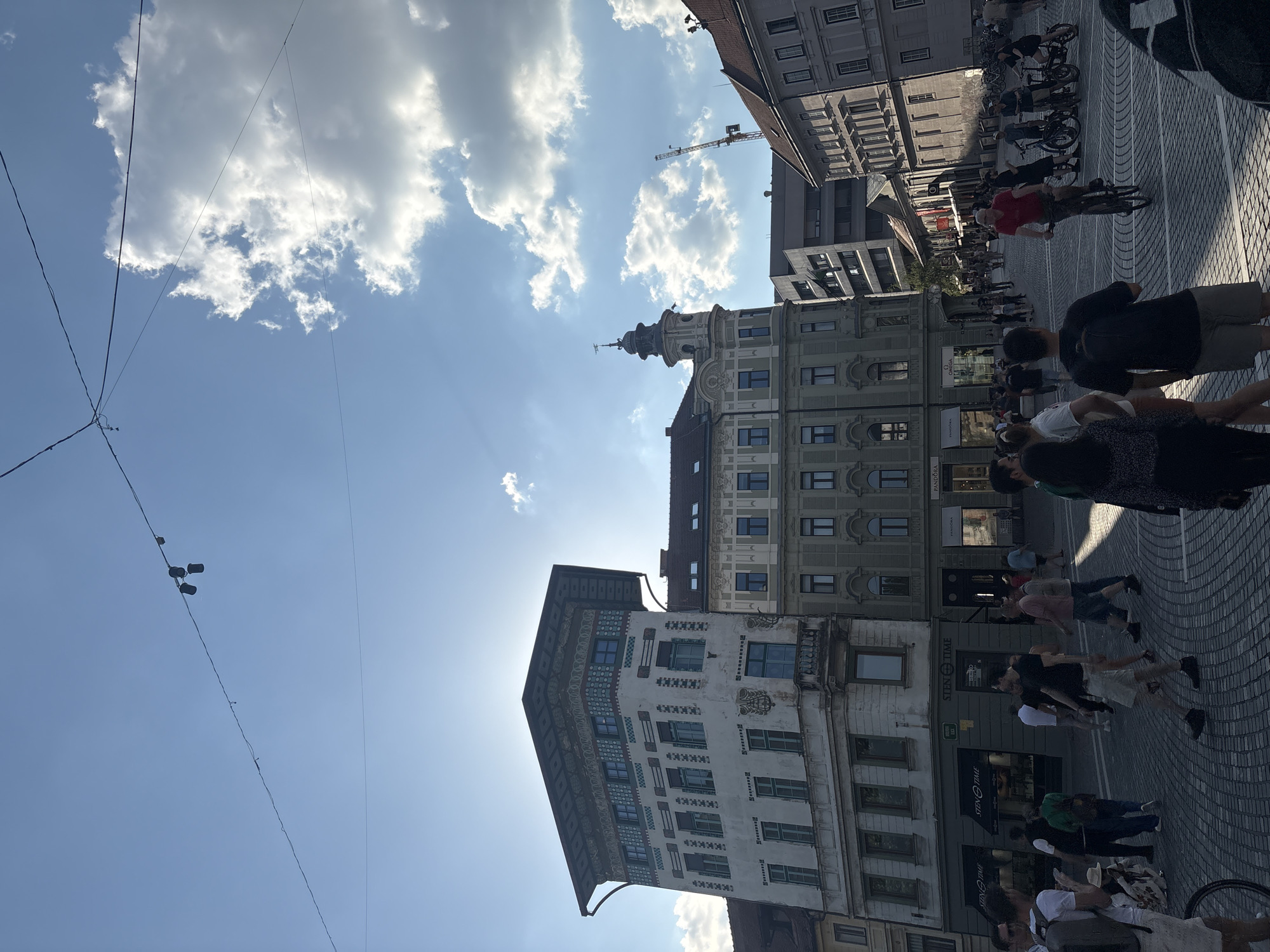 A street scene in a city square shows pedestrians walking on a cobblestone pavement under a bright sky with scattered clouds. The buildings lining the square exhibit varied architectural styles, with one building featuring a distinctive green-patterned facade and a unique roofline. Another prominent building has a domed tower. The sun is shining, casting shadows on the pavement. In the distance, a construction crane is visible. Several people are walking, some are standing, and one person is riding a bicycle. Bicycles are also parked in groups near some buildings. The overall impression is a lively urban environment on a sunny day.