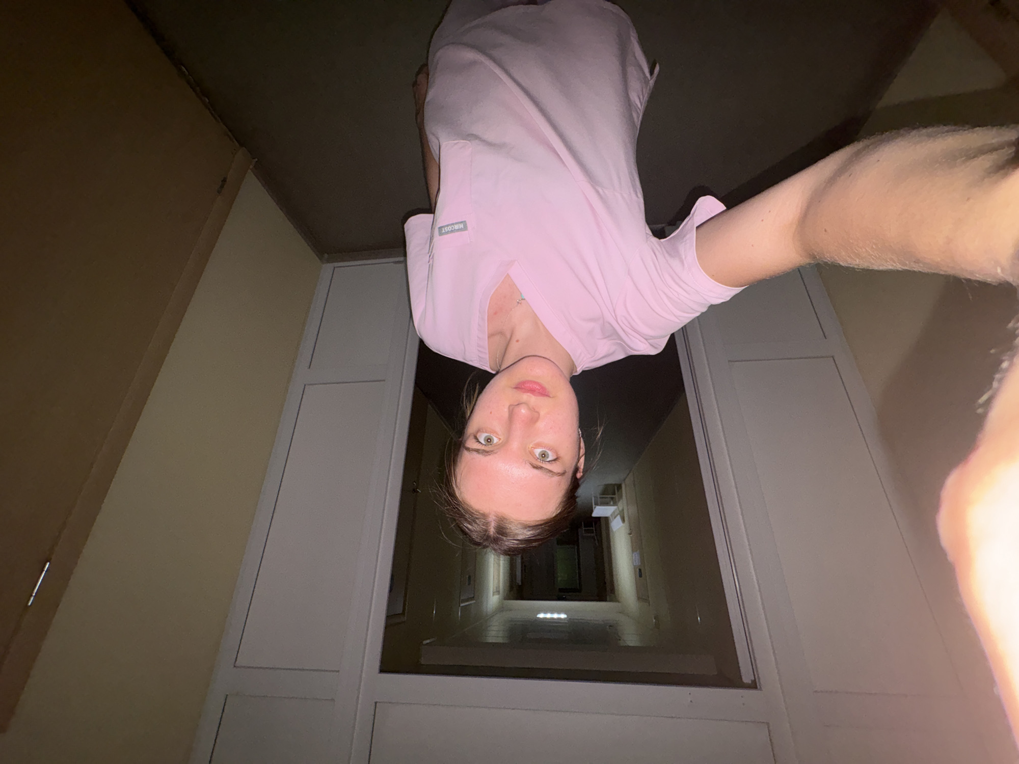 A woman in a pink scrub top is holding a camera above her head, pointed downward. The background is a long corridor in Perm, Russia. The corridor is framed by white doors and beige walls. There is a line of doorways going into the distance. There are florescent lights along the corridor.