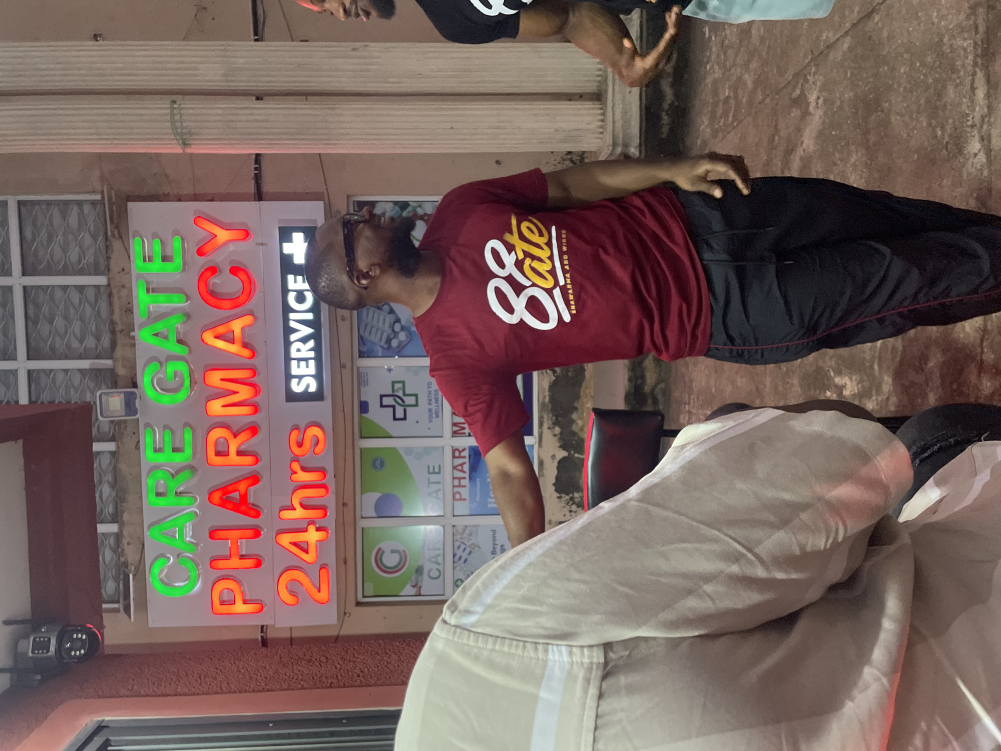The image shows a street scene at night outside a pharmacy in Abuja, Nigeria. A large, illuminated sign above the entrance reads "CARE GATE PHARMACY 24hrs SERVICE" in neon green and red letters. To the left, a security camera is mounted on the building's facade. In the foreground, three men are visible. One man, wearing glasses and a red t-shirt with the logo "8ate Shawarma and Wings", is standing with his left arm extended, seemingly gesturing or touching something just out of frame. He is wearing black track pants with a red stripe. Another man, partially visible on the right, is wearing a black t-shirt and appears to be engaged in conversation. A third person, whose body is mostly obscured by a beige garment, is on the far left, facing away from the camera. The environment appears to be paved, with a few structural elements like columns visible on the right. The lighting suggests it is nighttime, with the neon sign being the primary light source.