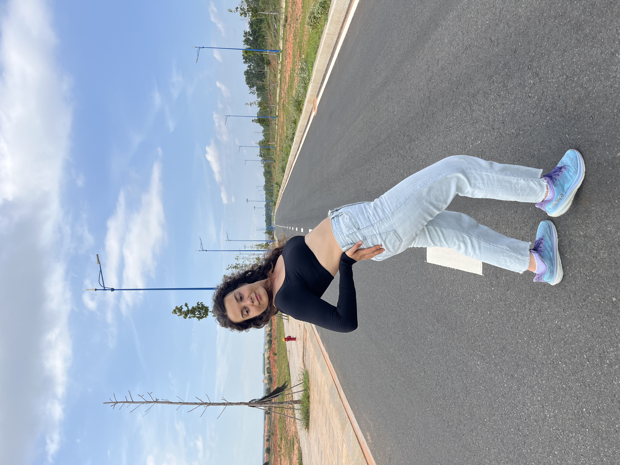 In Mui Ne, Vietnam, a young woman with curly dark hair is captured from the waist down, posing on an empty asphalt road. She is wearing a black long-sleeved crop top that exposes her midriff, light blue jeans, and bright blue and purple athletic shoes. Her body is arched backward, with her left hand resting on her hip and her right arm bent, creating a dynamic pose. The road stretches into the distance, flanked by grassy verges and scattered trees. The sky above is a clear blue, dotted with wispy white clouds. In the background, lampposts line the side of the road, and the landscape appears to be a developing area with construction visible in the distance. A red fire hydrant stands near a paved walkway on the left side of the road.