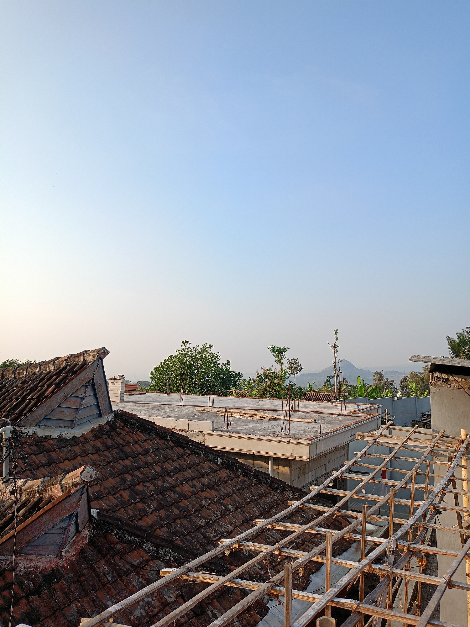 This is an outdoor, eye-level, full shot of a cityscape during the daytime in the city of Batu, Indonesia. The scene depicts rooftops with some under construction. In the foreground, there are weathered, reddish-brown tiled roofs, some with visible wooden beams and scaffolding suggesting ongoing construction or renovation. Adjacent to these, a concrete slab roof is visible, with rebar protruding from it, also indicating construction. In the middle ground, a cluster of green trees stands out against the sky. Further in the background, rolling hills and a mountain are discernible under a clear, pale blue sky. The lighting suggests either early morning or late afternoon sun. No people are visible in the frame.