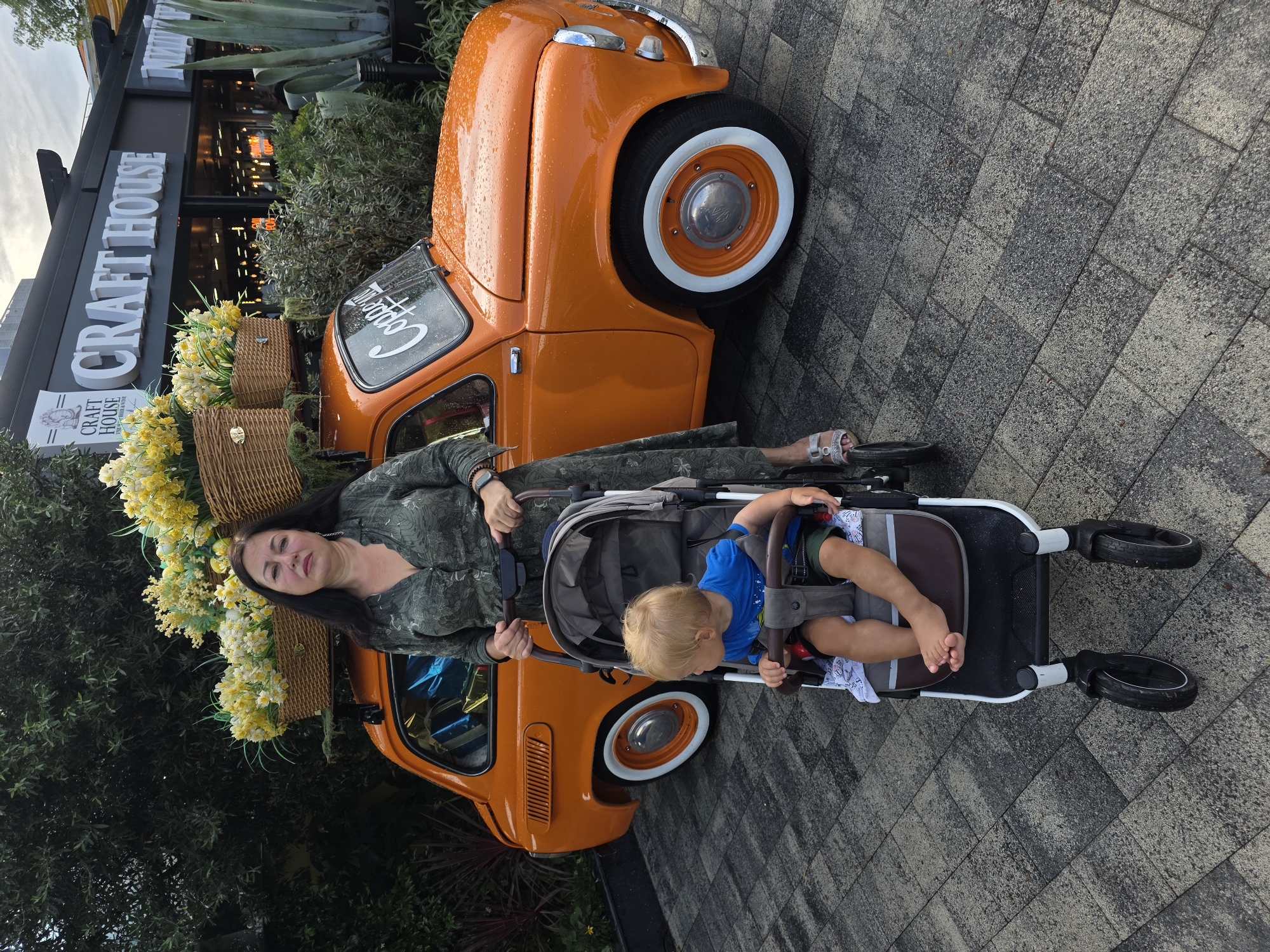 In Sochi, Russia, a woman stands next to an orange vintage car adorned with baskets of yellow and white flowers on its roof rack. She is holding the handle of a stroller, which contains a young blond child wearing a blue shirt and dark shorts. The car is parked on a paved area in front of a building with the sign "CRAFT HOUSE" in large white letters. The scene appears to be outdoors, with greenery visible in the background, and the car's surface shows droplets of water, suggesting it may have recently rained. The woman is wearing a patterned green dress and silver sandals.