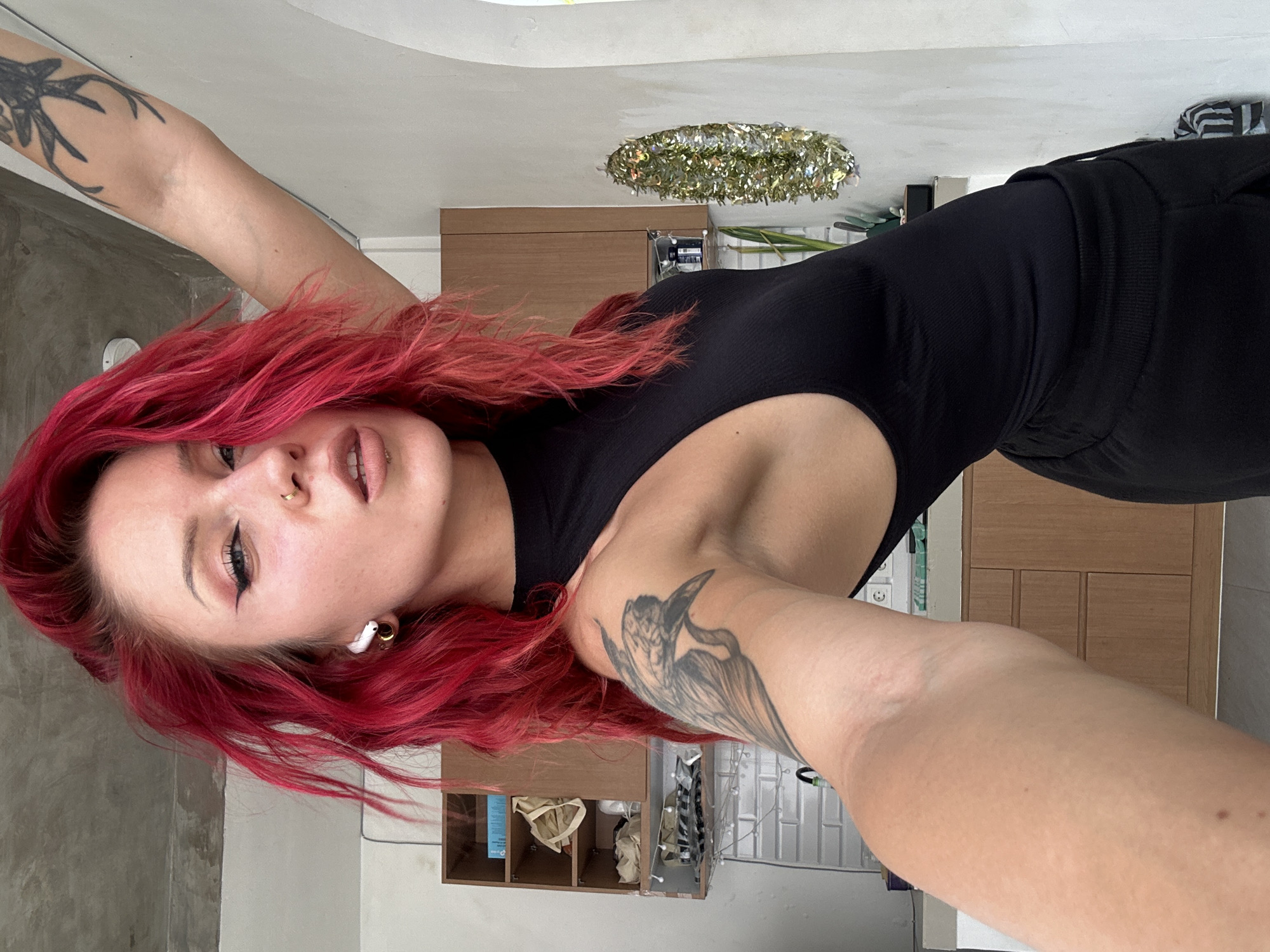 A woman with red hair is posing for a selfie. She is wearing a black top and has tattoos on both arms. The background includes a cabinet, a decorative light fixture, and a wall. The location is presumably indoors. The location in the city of North Kuta, Indonesia, is not discernible from the image.