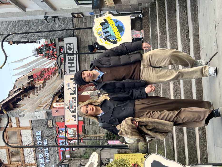 The image captures two individuals, a man and a woman, descending a stone staircase in an urban area identified as Yuzhong District, Chongqing, China. The woman, positioned on the left, has shoulder-length blonde hair and is dressed in a black double-breasted jacket over a light blue collared shirt, brown wide-leg trousers, and black shoes. She holds a light brown fur coat draped over her left arm. Her right hand is clasped with the man's left hand. The man, on the right, wears a black knit beanie, a black puffer jacket over a brown half-zip long-sleeved shirt, light brown relaxed-fit jeans, and light grey athletic shoes. Both individuals have neutral expressions, with the man looking slightly to his right.
The immediate foreground consists of worn, grey stone steps. At the bottom of the stairs, a grey paved surface is visible. To the right of the man, a white, rounded-top sign displays a graphic of a blue and green globe with a cityscape and the word "CHONGQING" in yellow text, resting on a stone pedestal.
The background features multi-story buildings constructed with grey stone and dark wood, exhibiting traditional Chinese architectural styles with tiered roofs and eaves. Multiple signs with Chinese characters are visible on the buildings, including "桐润亮·悦造型" and "问香茗茶". A prominent black sign displays "THE K2 BEAUTY" in white lettering. Overhead, white fabric banners are suspended across the street, beneath which hang a series of circular, tiered lanterns or decorations. Further in the background, to the upper right, a decorated Christmas tree adorned with red elements, possibly red envelopes, is visible on a rooftop or balcony. Another individual in dark clothing is partially visible further up the staircase in the background. The scene is illuminated by natural daylight.