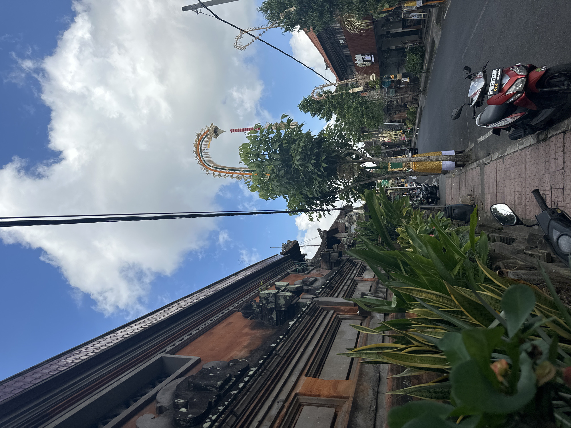 The image depicts a street scene in the Ubud District, Indonesia, on a partly cloudy day. On the left, a traditional Balinese building with intricate stone carvings and reddish-orange brickwork lines the sidewalk. Lush green plants, including snake plants, grow along the base of the wall. The sidewalk itself is paved with red bricks.
The street is paved and appears to be moderately busy with parked motorcycles and a few people in the distance. A red and silver scooter is prominently parked on the right side of the sidewalk. Several trees line the street, some adorned with decorative, arched structures that appear to be ceremonial or festive decorations. These decorations, made of woven materials and possibly flowers, are a common sight during Balinese festivals. Buildings with traditional architecture and various shops are visible on the right side of the street. Overhead, power lines crisscross the sky, and fluffy white clouds are scattered against a bright blue sky. The overall impression is a vibrant street in a culturally rich area, likely during a festive period.