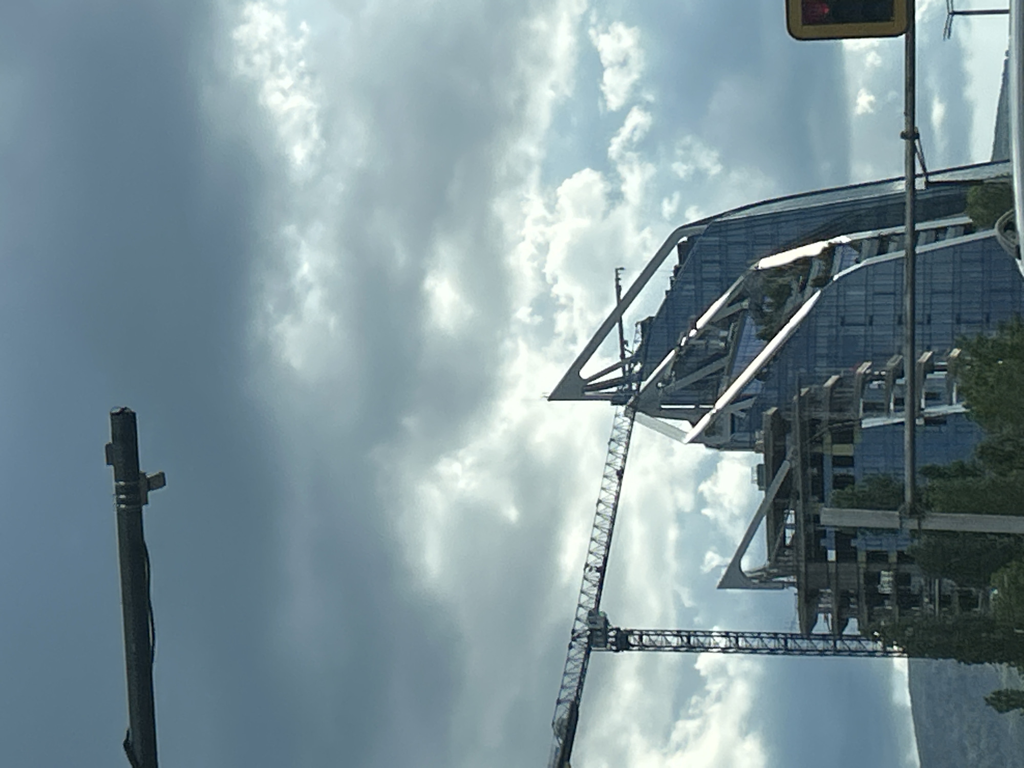 The image shows a construction site in Sofia, Bulgaria. Two construction cranes are prominent against a cloudy sky. A partially constructed building with a steel framework and glass façade is visible. In the upper right, a traffic light is visible. A utility pole is on the left.