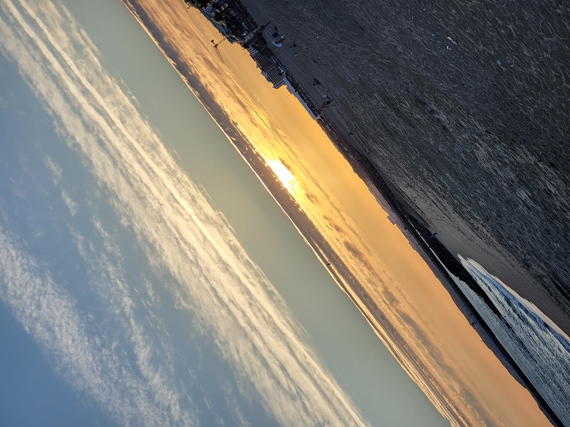 The image depicts a beach in Quarteira, Portugal, at sunset. The horizon line is tilted, with the sun low in the sky, casting a warm orange and yellow glow across the clouds and the sea. The foreground is dominated by sand, textured with ripples from the receding tide, and scattered with a few small shells. A person can be seen walking along the water's edge in the distance. Further inland, the beach is populated by a few individuals, some sitting and others standing, with buildings and a construction crane visible in the background. The sea is calm, with small waves lapping against the shore. The sky above is a gradient of blue, transitioning into the warm sunset hues.
