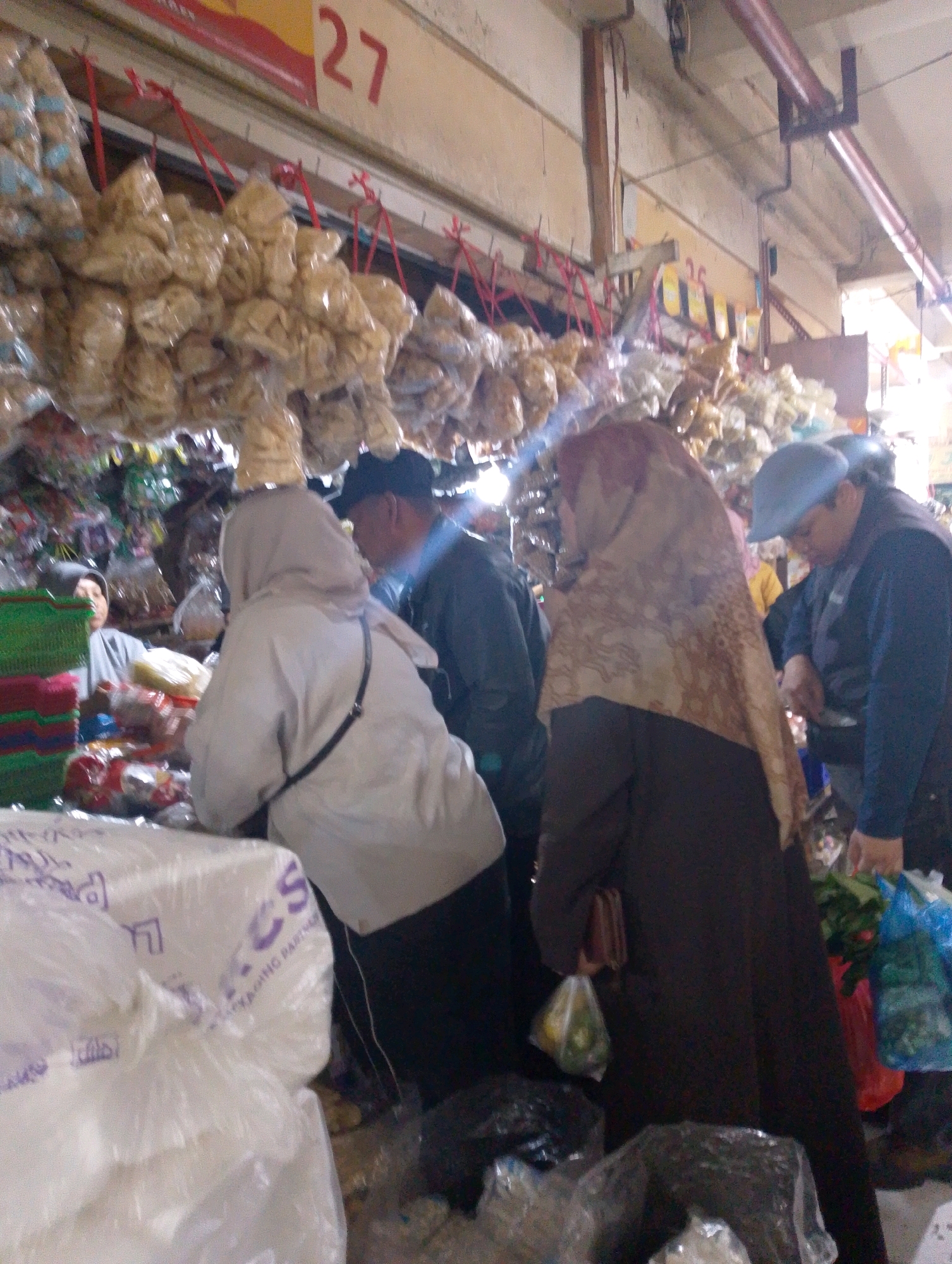 The image depicts a market scene in Lembang, Indonesia. In the foreground, large plastic-wrapped items are piled up. In the mid-ground, several people are interacting in what appears to be a food stall. A woman wearing a grey hijab and a white garment is interacting with another person. To her right, a man in a black cap and dark jacket is looking down. Further to the right, a woman in a brown hijab and dark dress is holding a small purse and a plastic bag containing produce. She is standing next to a man wearing a blue cap and dark jacket, who is also looking down. Hanging from the ceiling are numerous bags of dried goods or snacks. In the background, a sign with the number "27" is visible. The overall environment suggests an outdoor or semi-outdoor market setting with goods displayed for sale.