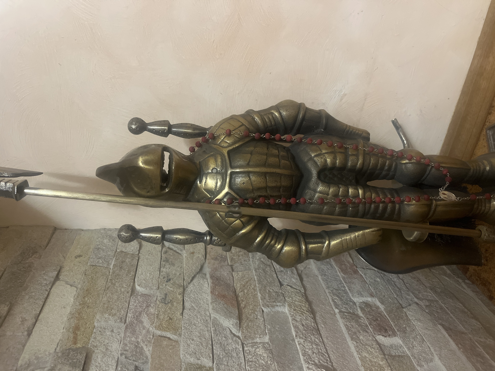 The image shows a statue of a knight in armor, possibly a decorative piece for a fireplace or a mantelpiece. The knight is made of a metallic material, likely bronze or brass, with a golden sheen. It is holding a long pole or spear. A string of red beads, resembling rosary beads, is draped around the knight's neck. The knight is positioned in a corner, with a stone wall on the left and a light-colored wall on the right. There is a wooden floor or base at the bottom. The lighting suggests an indoor setting. The image does not contain any information that would place it in the city of Tarnivtsi, Ukraine, or any other specific location.