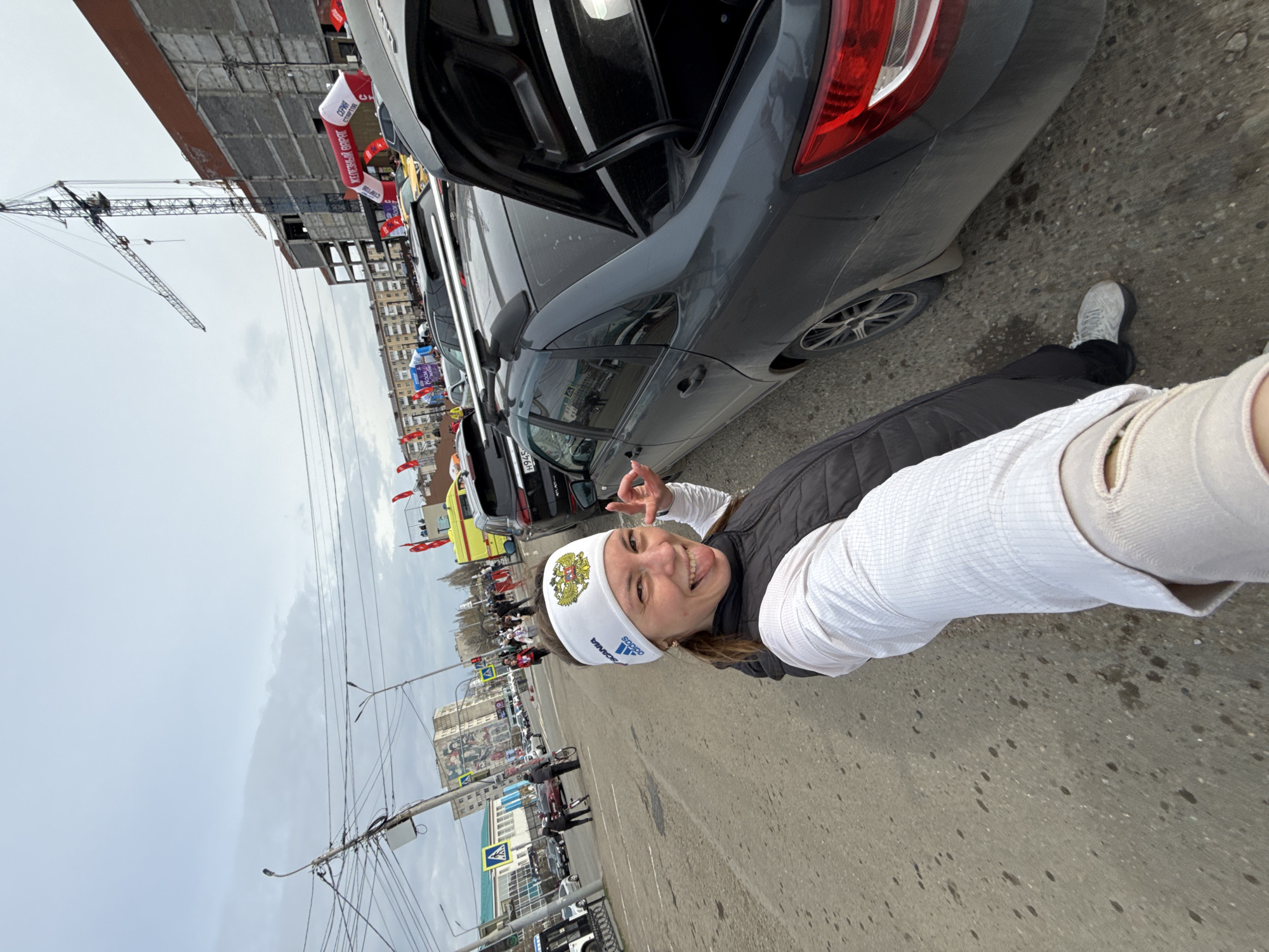 A woman smiles for a selfie on a street in Neftekamsk, Russia. She wears a white Adidas headband with the Russian coat of arms, a white long-sleeved shirt, and a black vest. She makes a peace sign with her left hand. Parked behind her is a gray sedan with its trunk open. Other parked vehicles, including a yellow ambulance, are visible in the background. A crowd of people and flags line the street further in the background. In the distance are buildings, street lights, traffic signs and a construction crane. The sky is overcast.