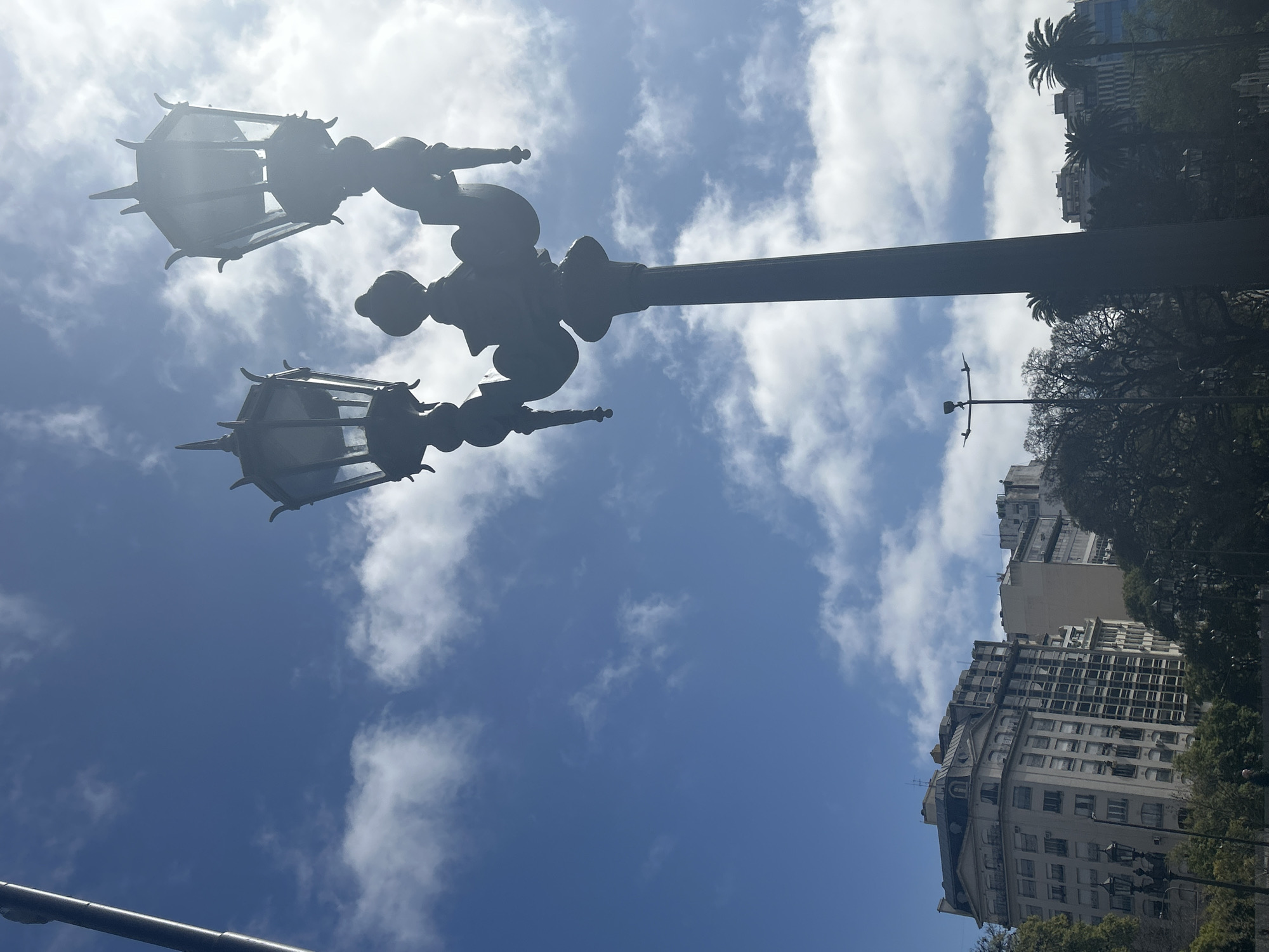 The image is a low-angle shot of a vintage double-headed lamppost against a bright blue sky with scattered white clouds. The lamppost is dark and ornate, with pointed finials on top of each lamp housing. Sunlight glints off the glass of the lamp on the right. In the background, to the left, are several multistory buildings, one appearing older with a mansard roof and decorative facade, and others more modern with rows of windows. Trees are visible in the middle and foreground, partially obscuring the lower portions of the buildings. The scene appears to be in an urban park or plaza. This view is consistent with the area around the Obelisco in Buenos Aires, Argentina, where similar lampposts are found lining avenues and public spaces. There are no people clearly visible in the frame.