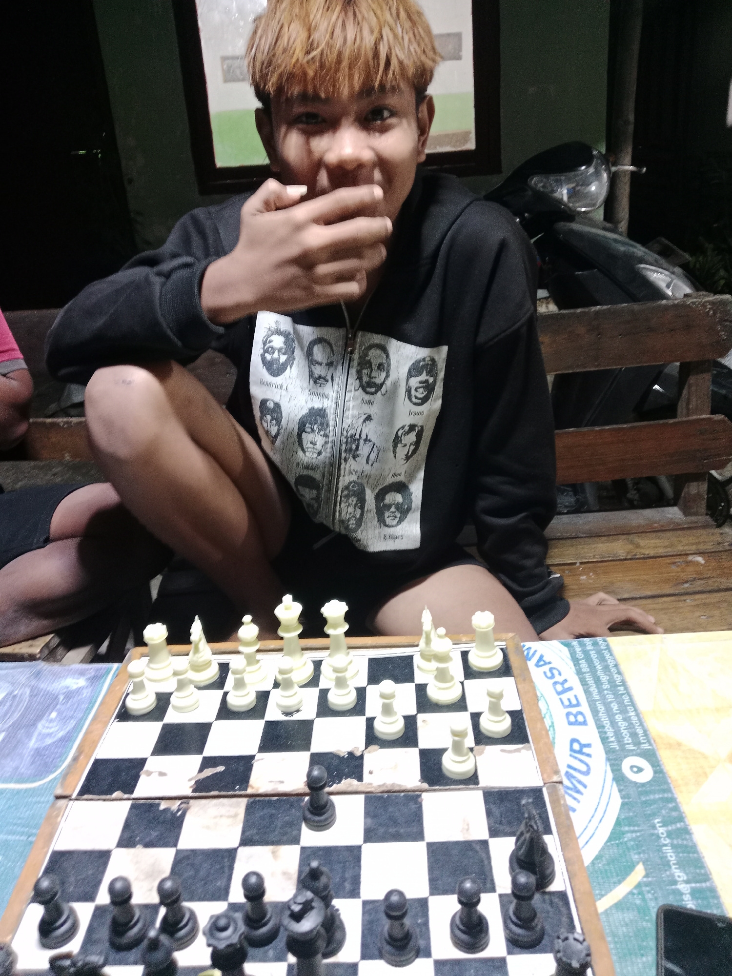 A young person with blonde hair sits on a wooden bench, playing chess at a table in Glagah Wangi, Indonesia. They wear a black hoodie with a graphic print of several faces. The chessboard is set up with white and black pieces. The scene appears to be outdoors in the evening, with a motorcycle visible in the background.