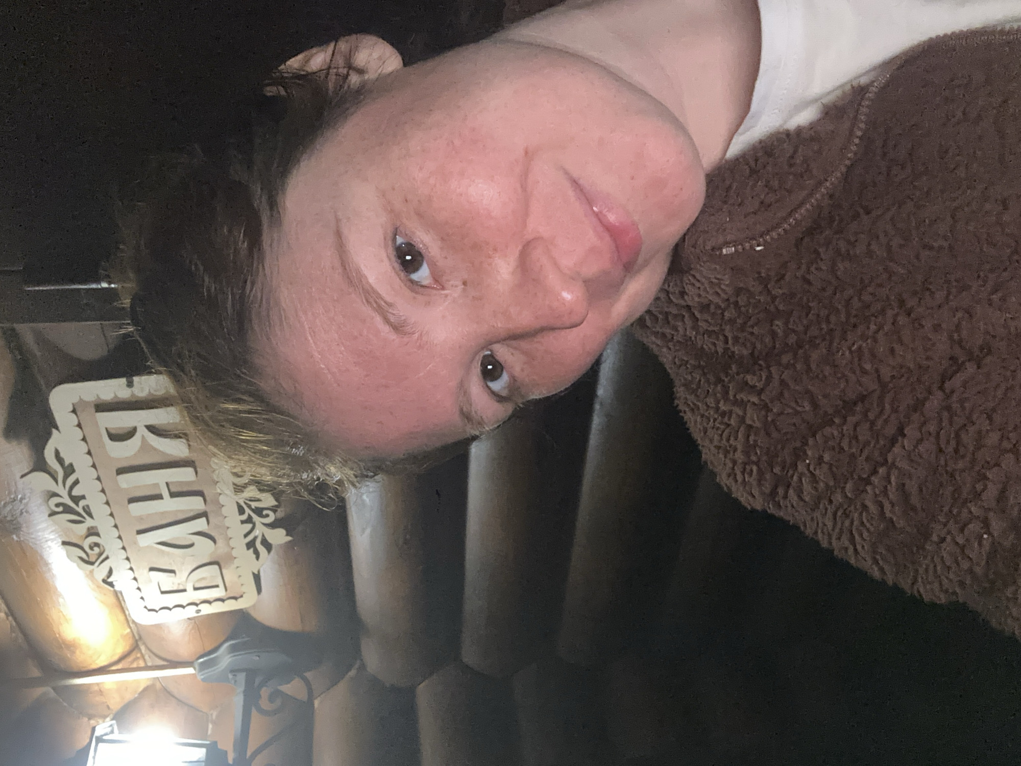 A woman is in a sauna. The background features light-colored, unfinished wood walls. A white sign with decorative filigree has the Russian word "БАНИ" on it, which translates to "Bathhouses". She is wearing a brown, textured top over a light-colored shirt. Based on the sign, this photo was likely taken in a bathhouse in Nizhny Novgorod, Russia.
